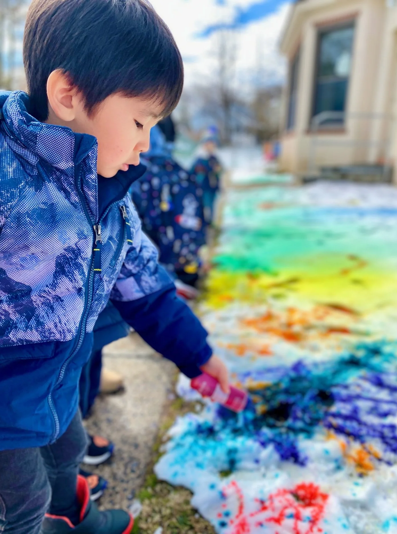 A young boy in a blue jacket painting on a large outdoor canvas with colorful splashes of paint. Other children are visible in the background, also engaged in painting. The setting appears to be outside a house on a bright day.