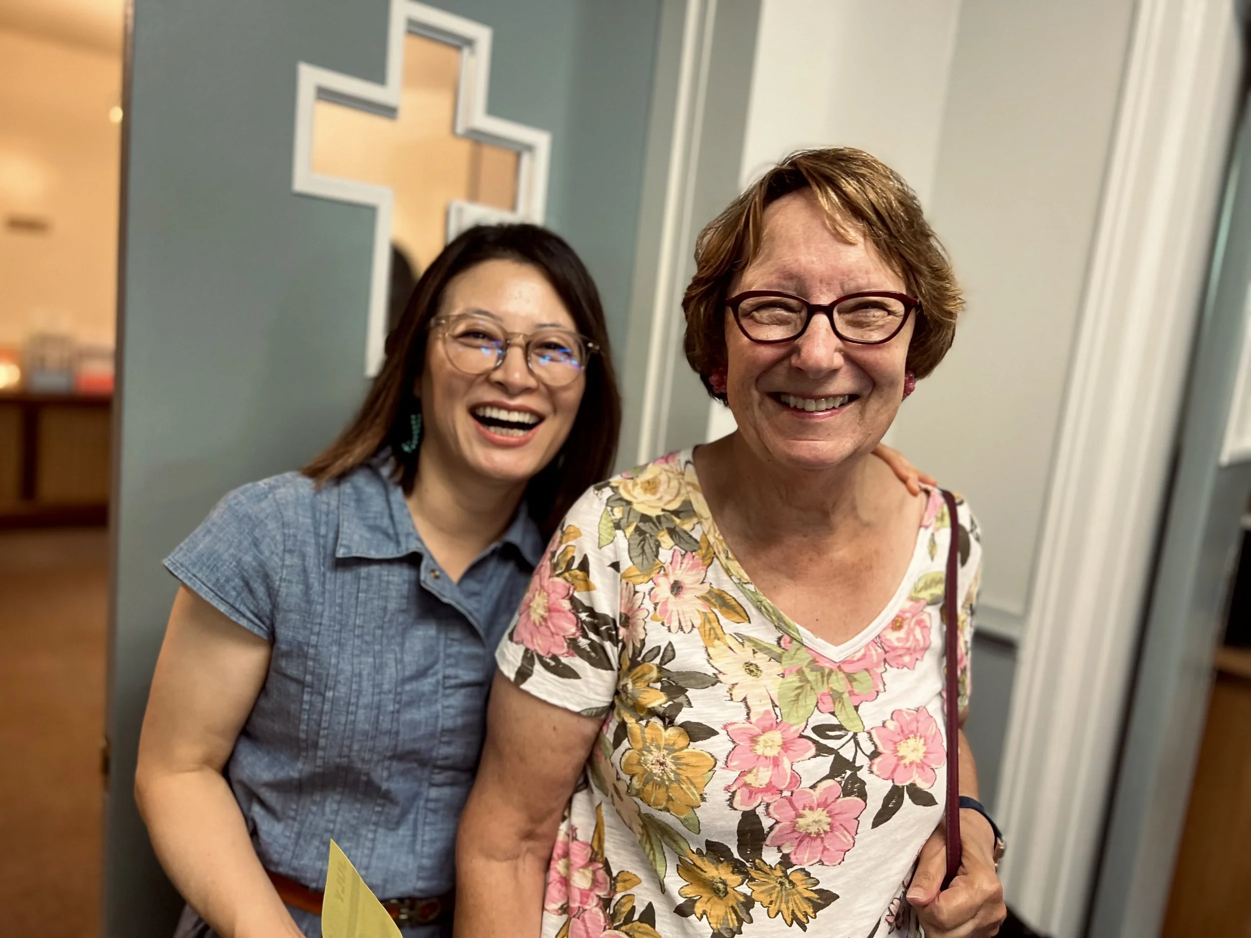 Two women smiling and posing together indoors, one older woman with glasses, a floral shirt, and a purse, and a younger woman with glasses, a blue shirt, and earrings, standing behind her.