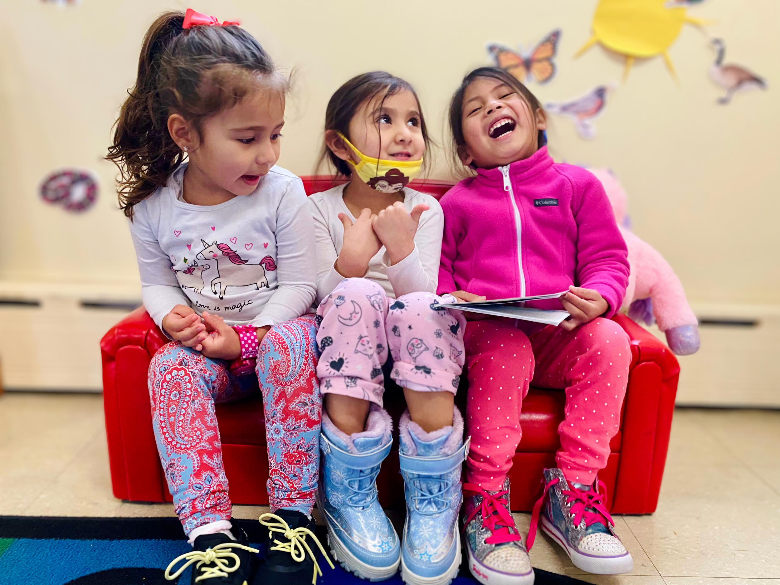 Three young girls sitting on a red couch, laughing and enjoying each other's company in a decorated indoor space.