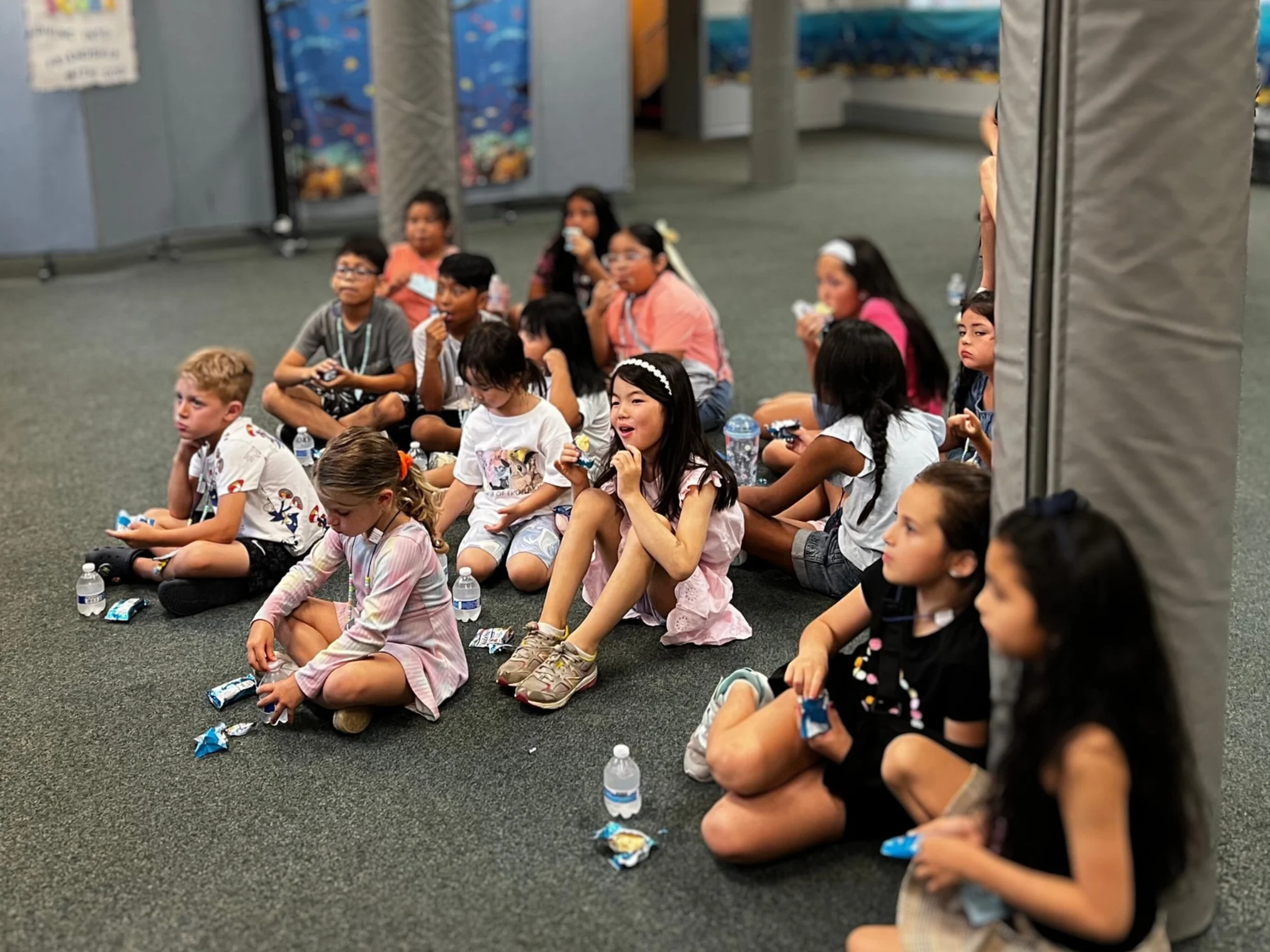 A group of children sitting on a carpeted floor in a room, possibly at a school or library, eating snacks and drinking bottled water.