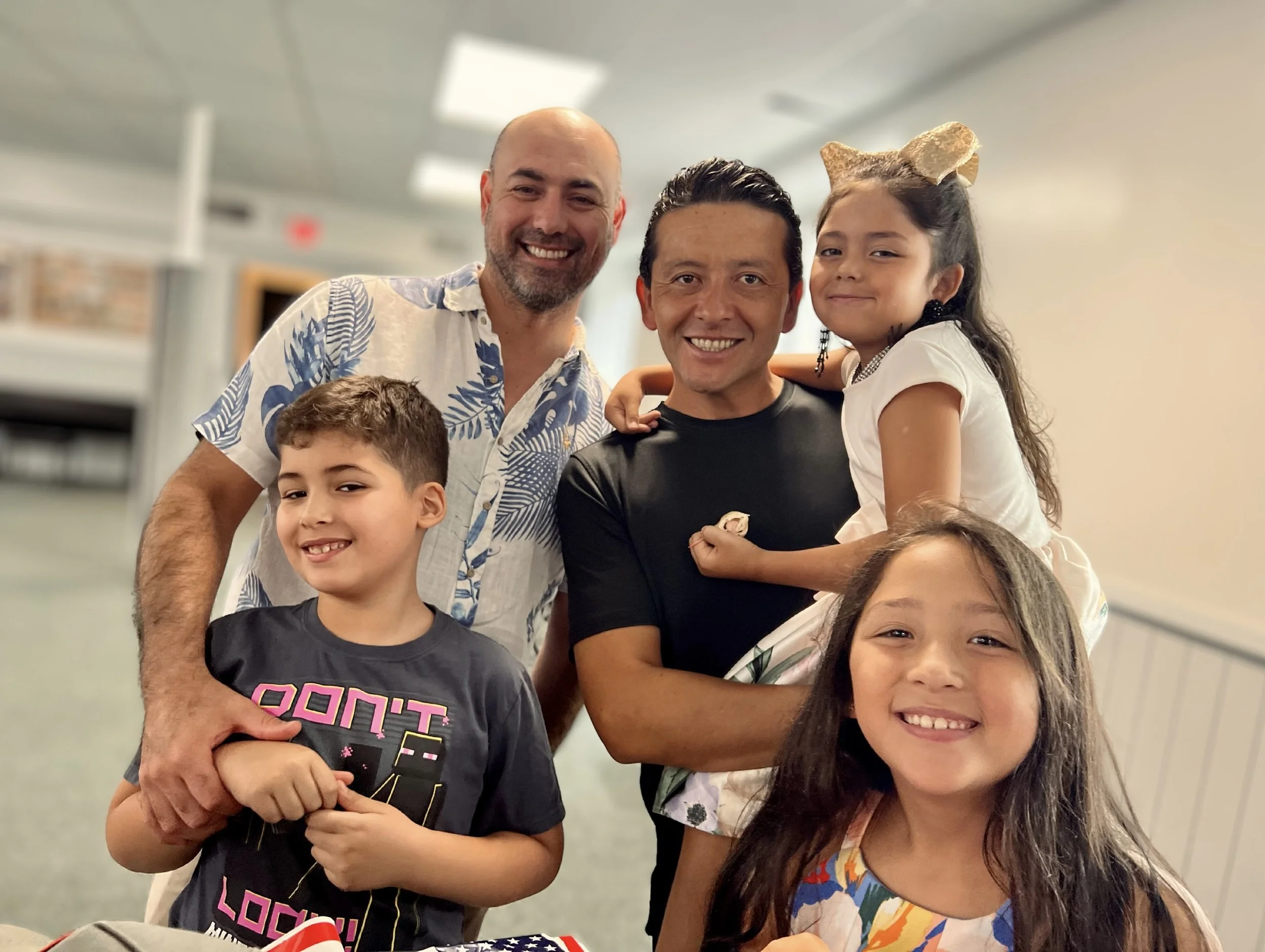 A group of four smiling adults and children posing together indoors, with two adults and three children, one girl being held by a man, in a well-lit room.