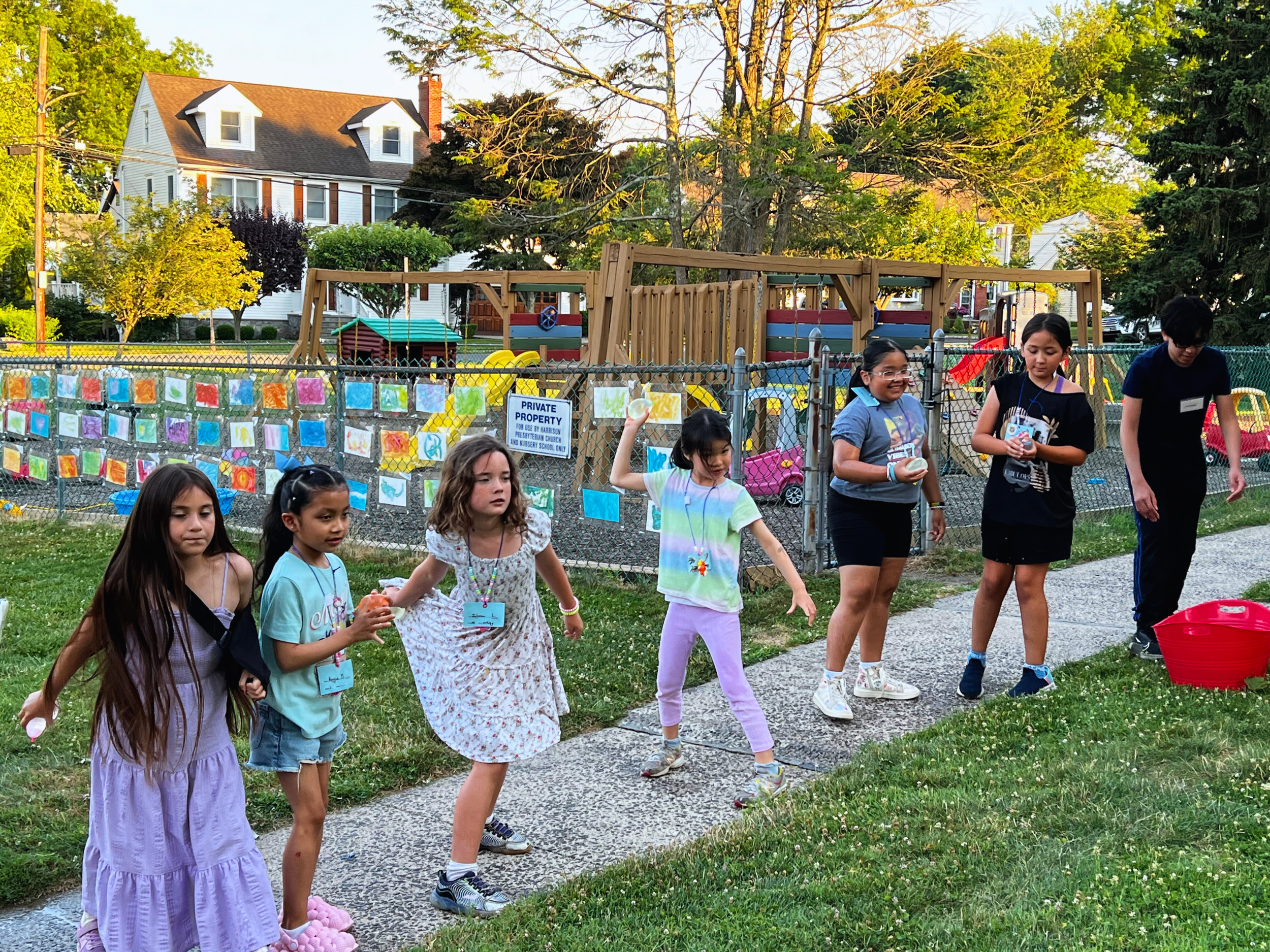 Children playing outdoors on a sidewalk in front of a fenced playground, with colorful artwork displayed on the fence and a house with trees in the background.