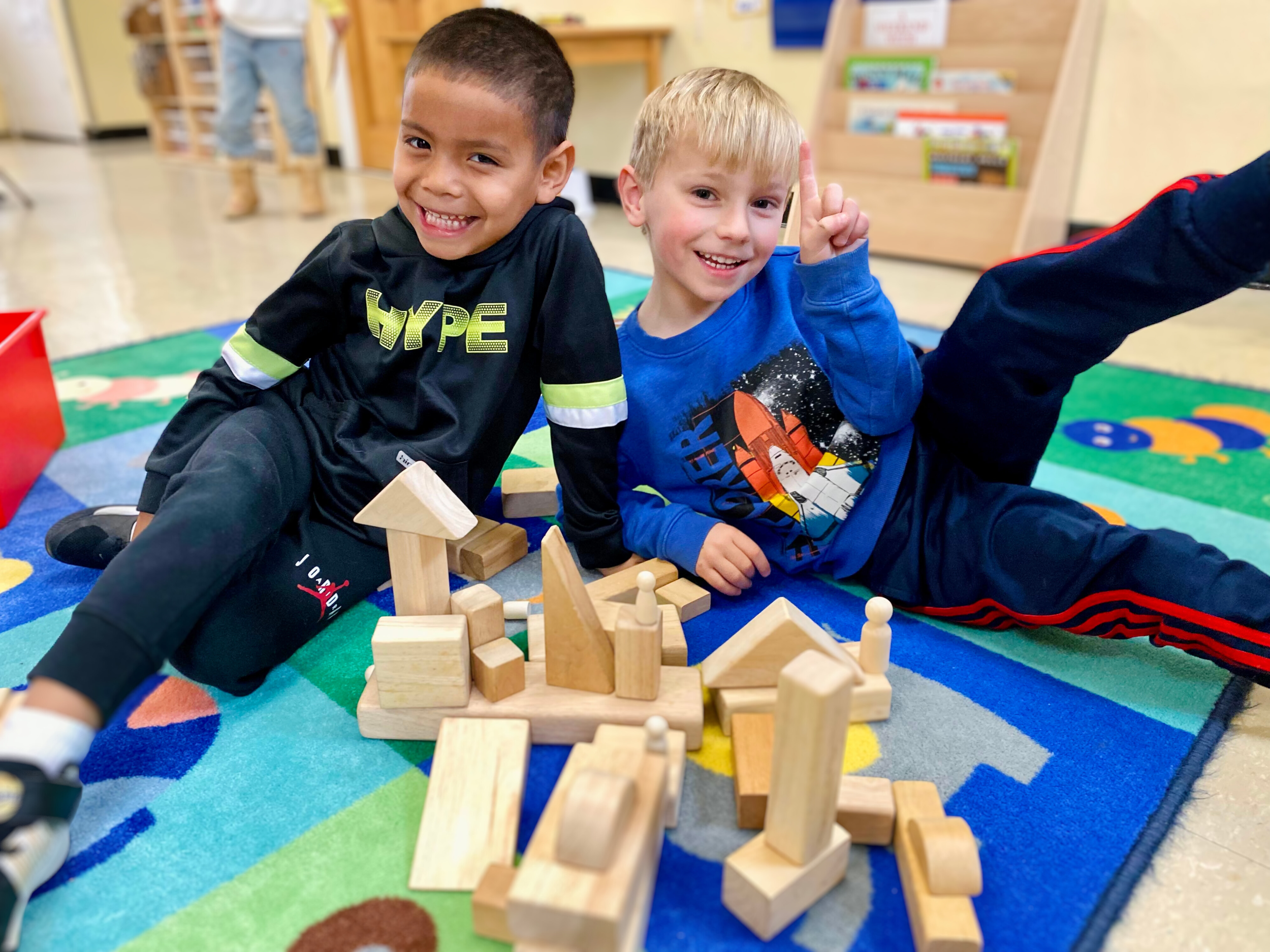 Two young boys smiling and playing with wooden building blocks on a colorful rug in a classroom or daycare setting.