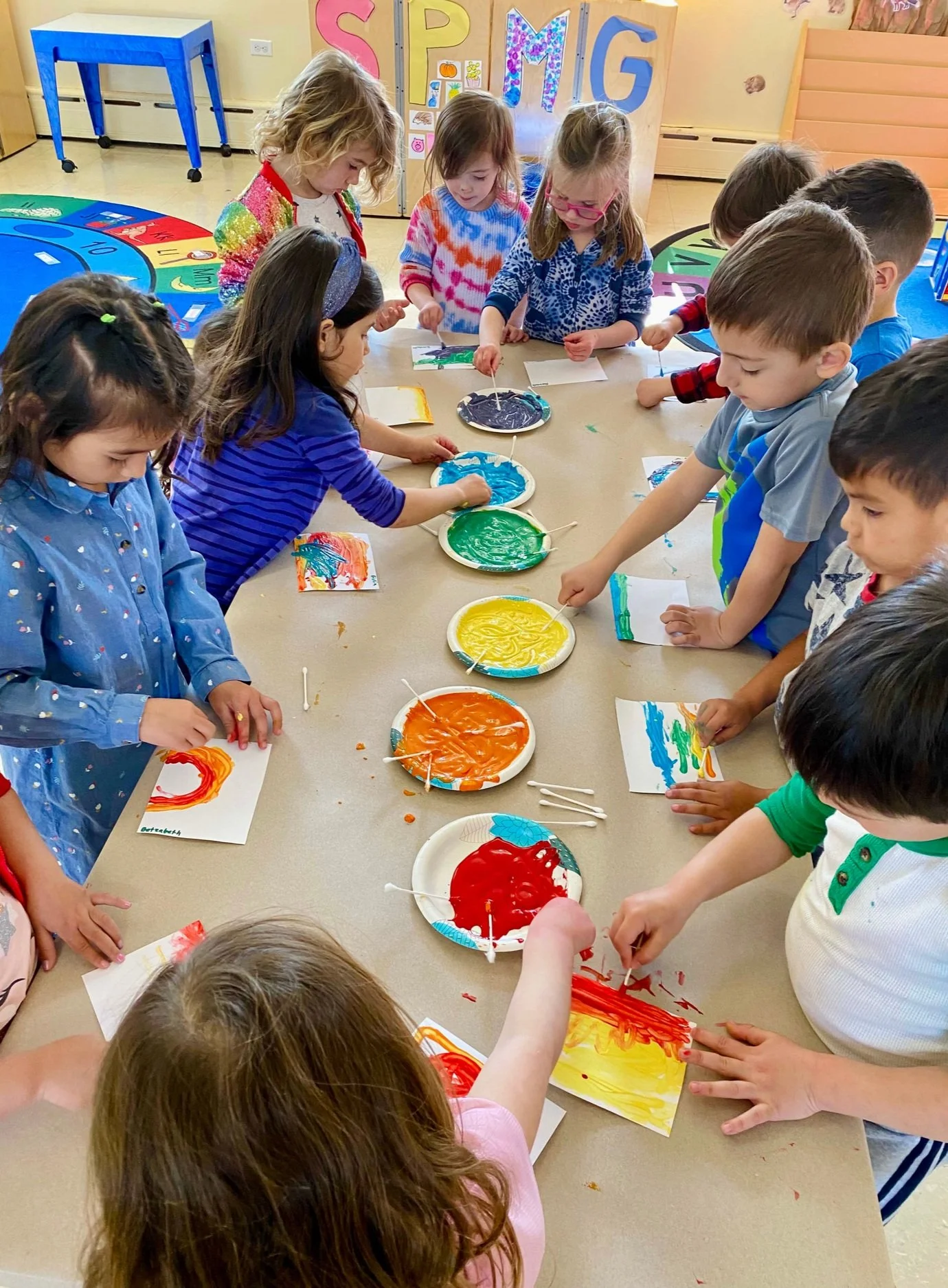 Children painting with colorful paints on paper at a table in a classroom.