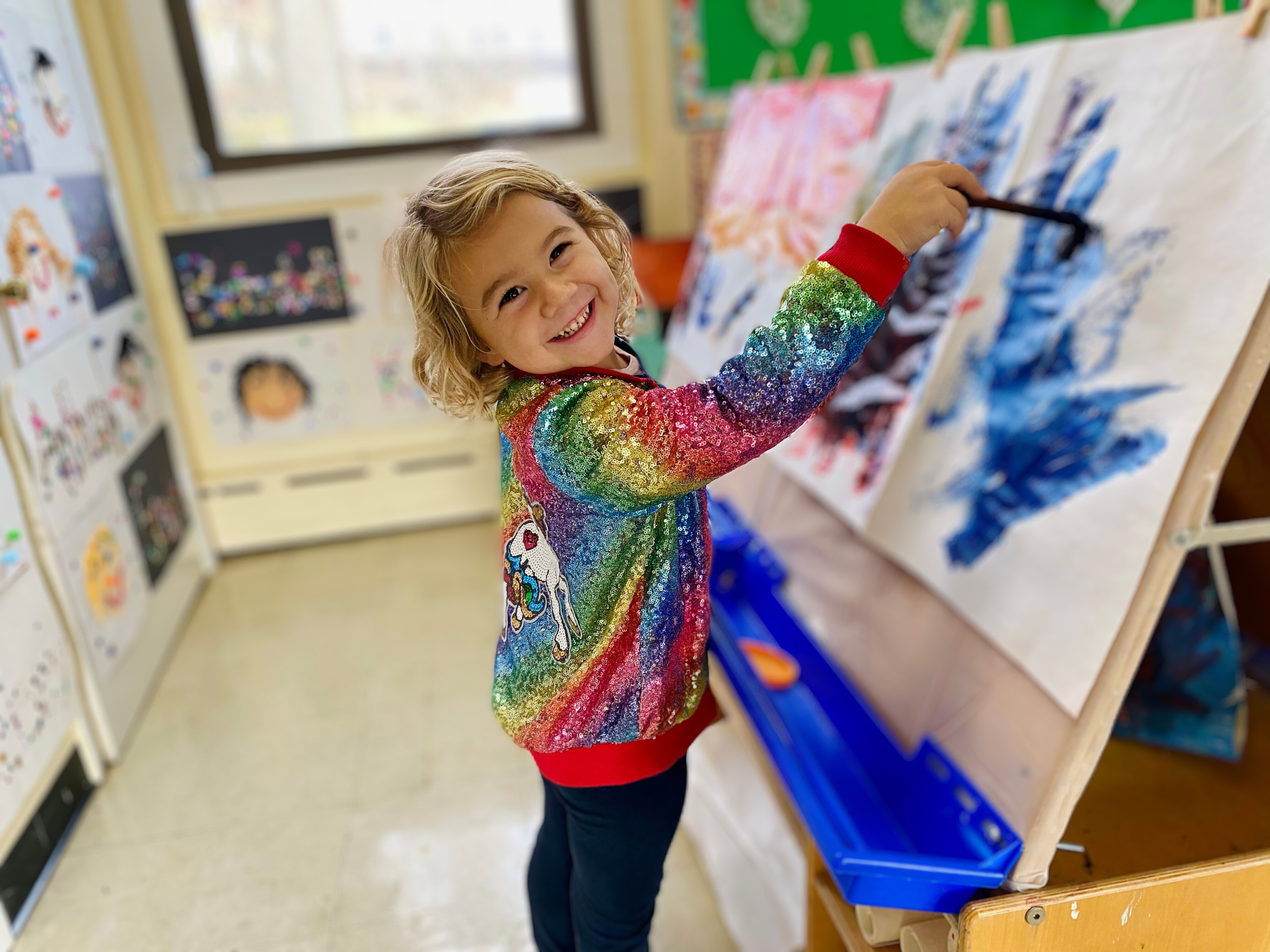 A smiling young girl with curly blonde hair in a rainbow-colored sequined unicorn shirt, painting on a large canvas in an art classroom.