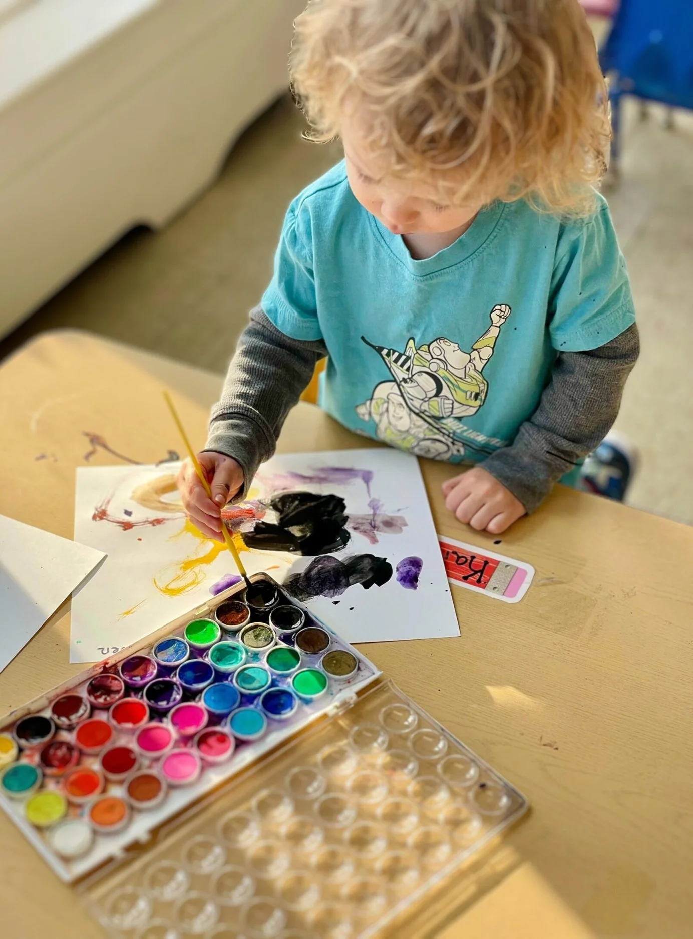 A young child with curly blonde hair painting with watercolors at a table. The child is wearing a blue t-shirt with a cartoon character and has a palette of various watercolor paints in front of them.