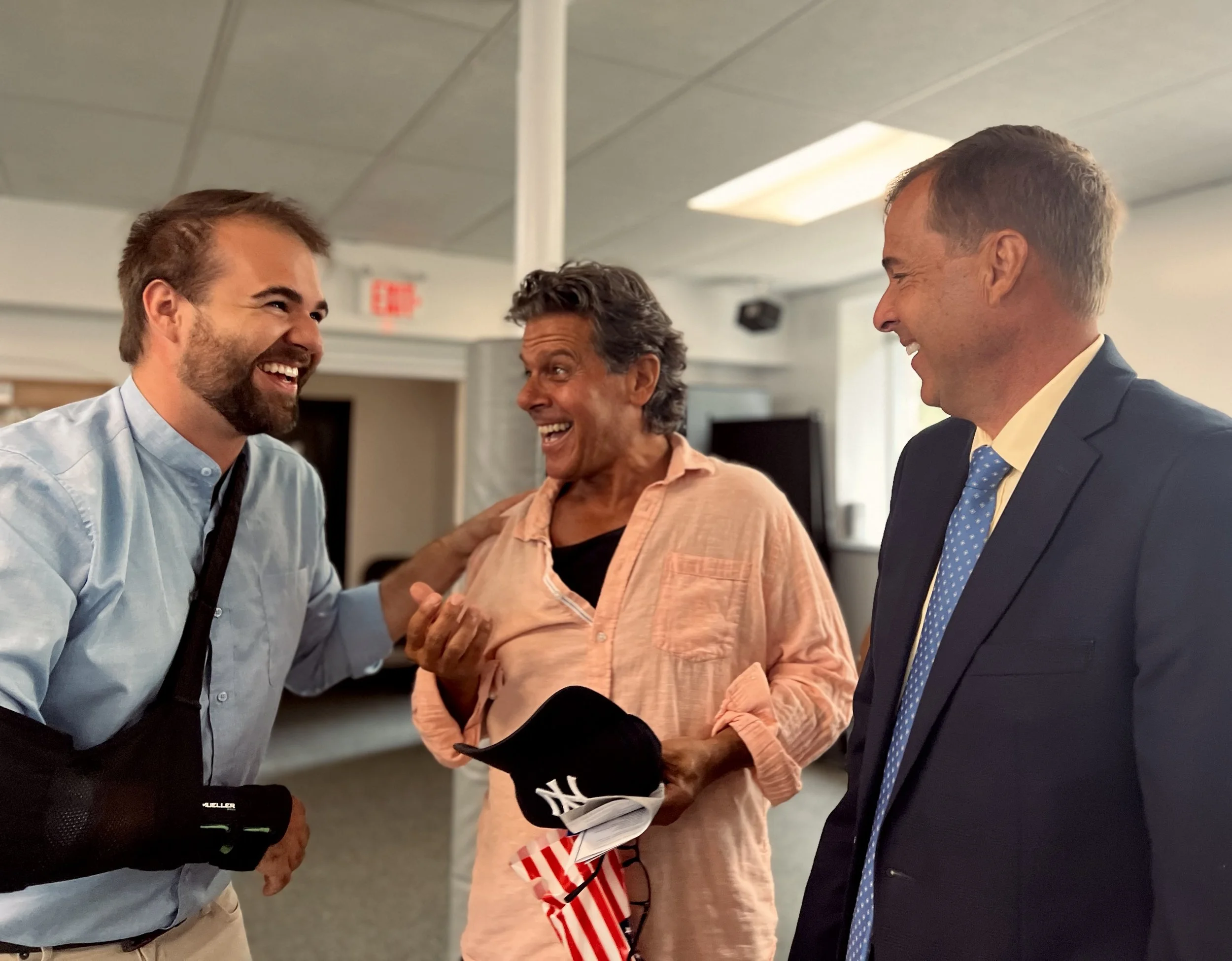 Three men smiling and talking in an indoor setting, with one man wearing a light blue shirt and a black arm brace, another man in a suit and blue tie, and a woman with short curly hair wearing an orange shirt holding a Yankees cap and an American flag. All appear happy and engaged.