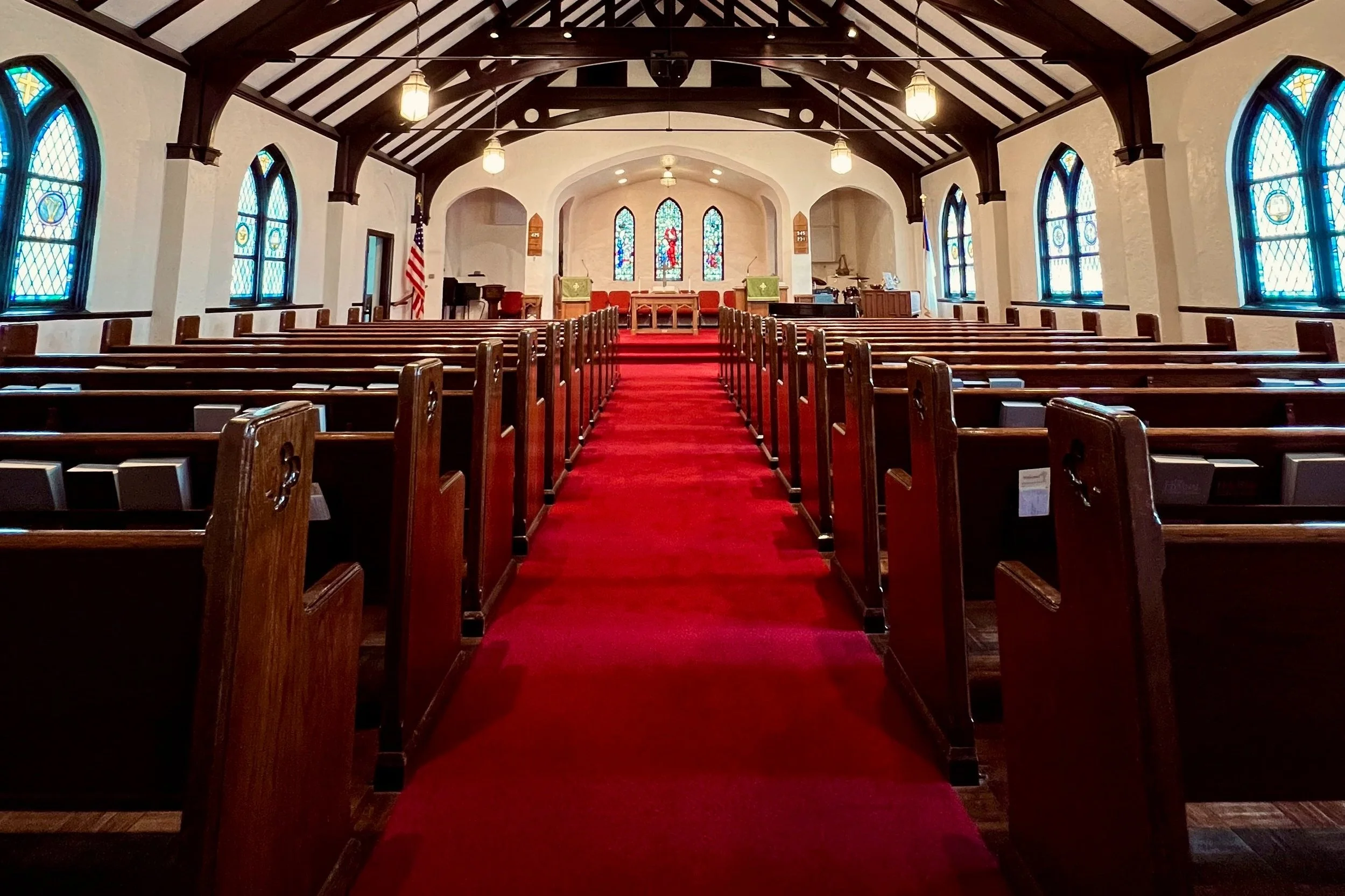 Interior of a church with wooden pews, stained glass windows, a red aisle runner, and an altar at the front with stained glass windows behind it.