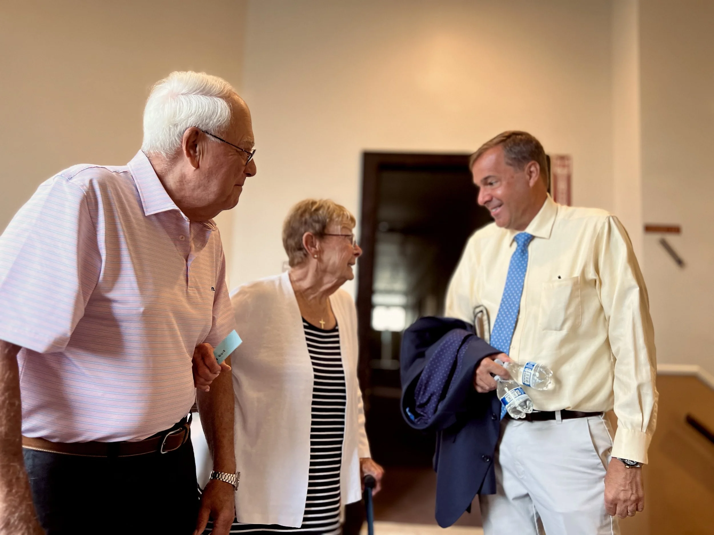 Four people, two men and two women, are standing and talking indoors. One man is holding two bottles of water, and the woman in the middle is using a cane. They are engaged in conversation and smiling.