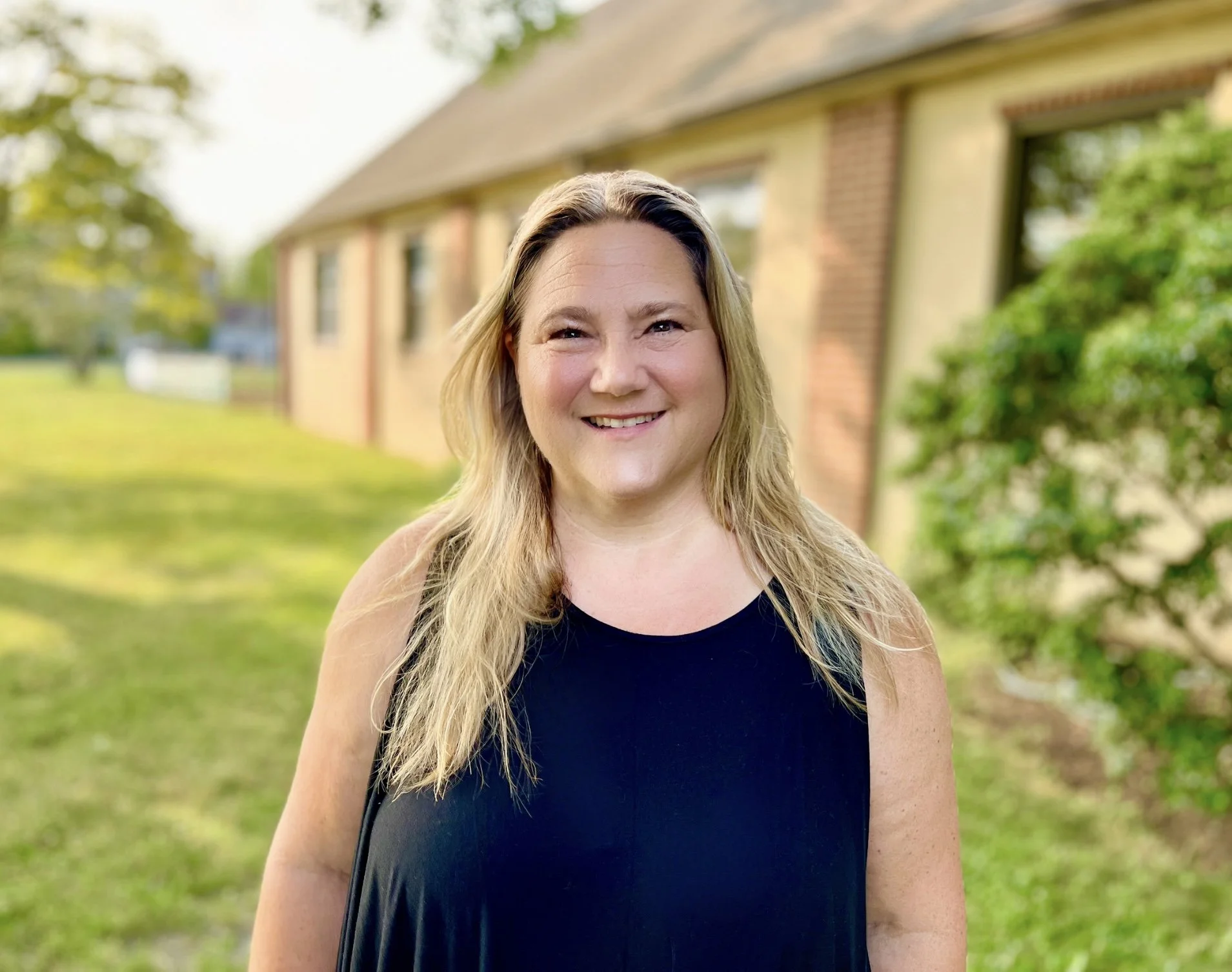 A smiling woman standing outside in front of a house with a lawn and trees.