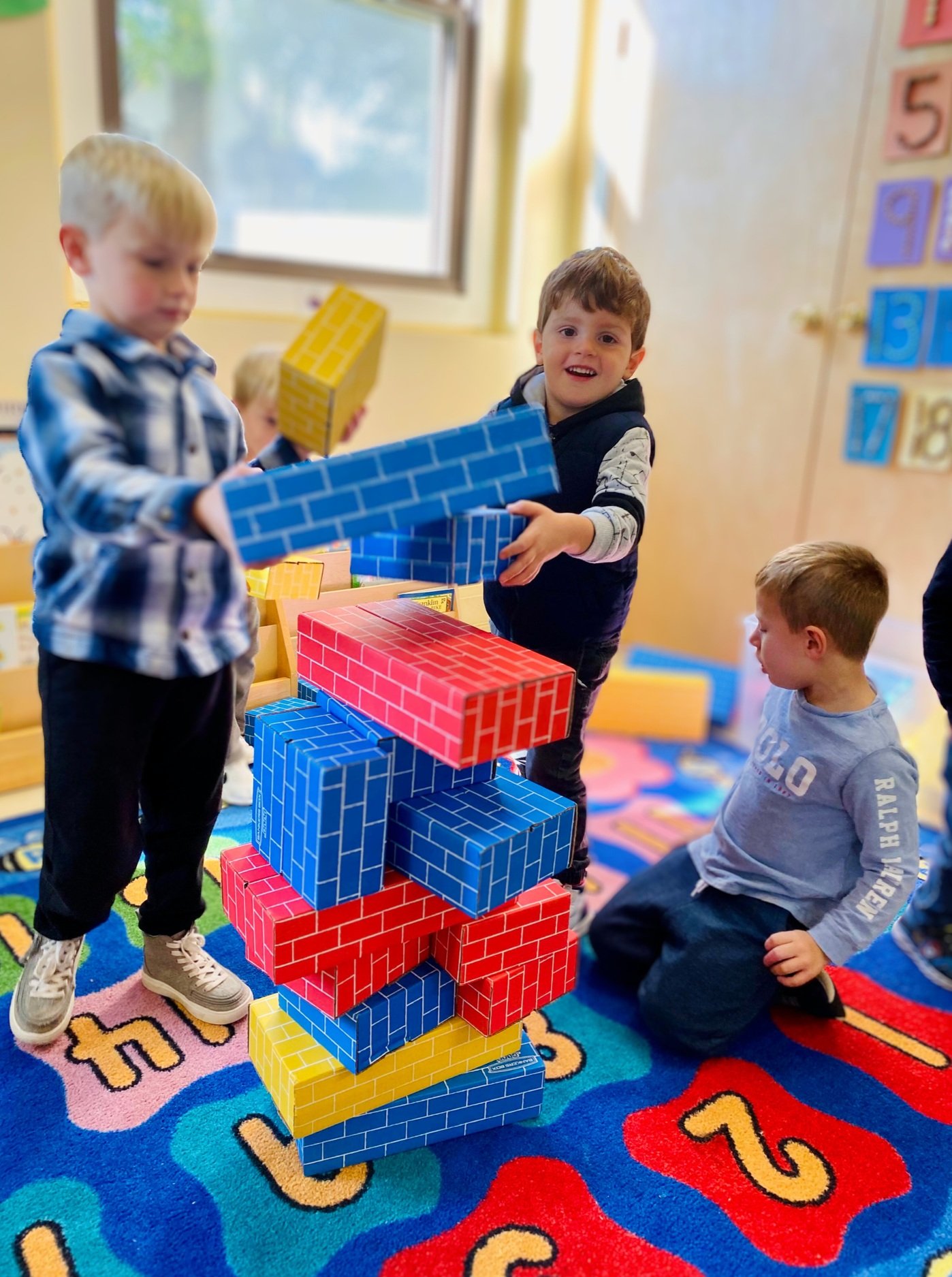 Children playing with large, colorful paper brick blocks in a classroom, building a tower on a vibrant, alphabet-themed rug.