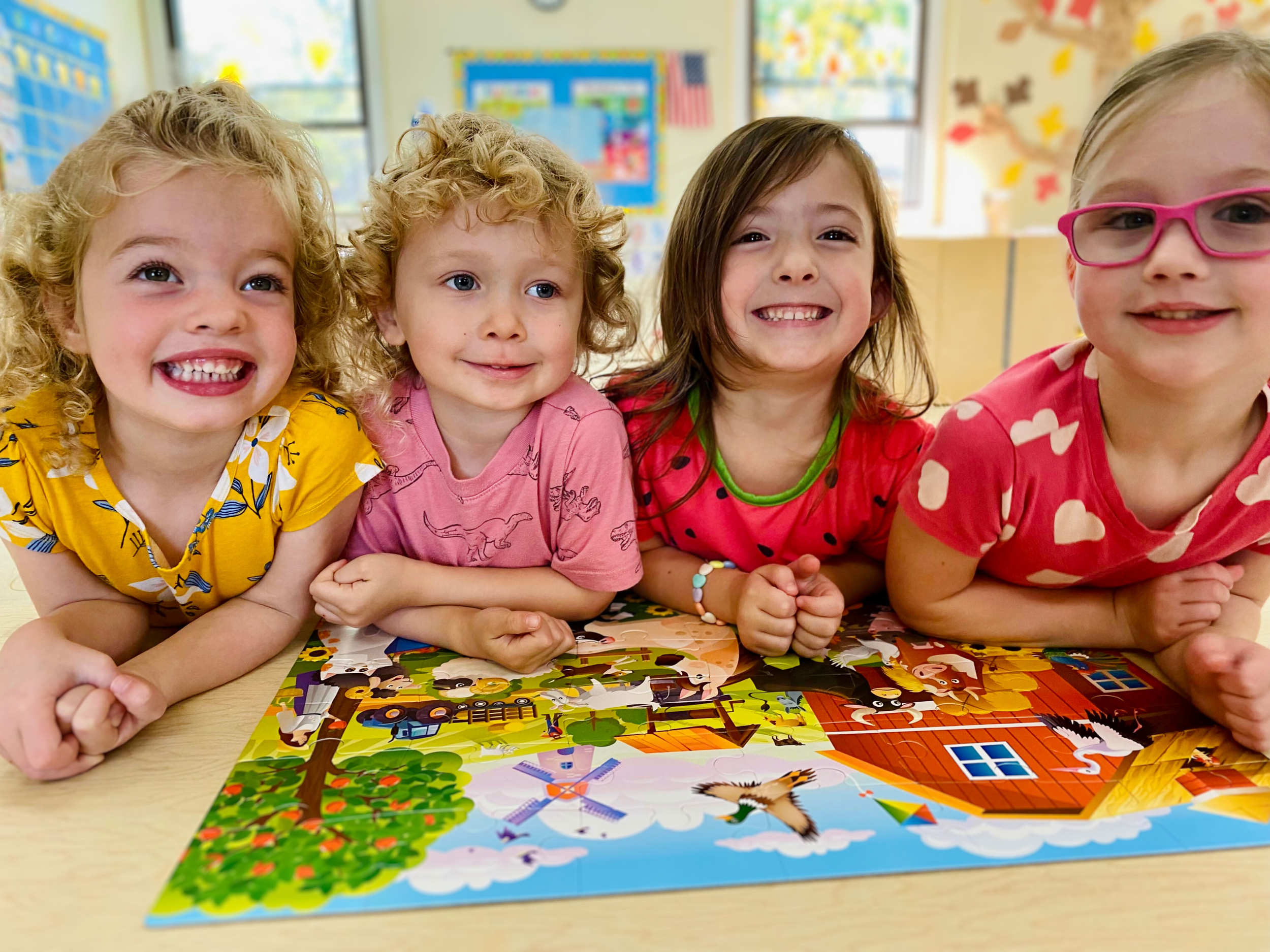 Four young girls smiling and lying on their stomachs around a colorful puzzle on a table in a classroom.