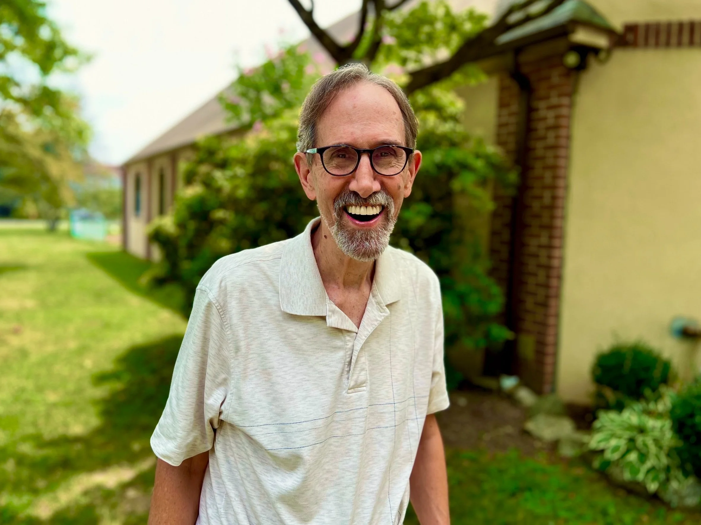 A smiling elderly man with glasses and a beard standing outside in front of a house and greenery on a bright day.