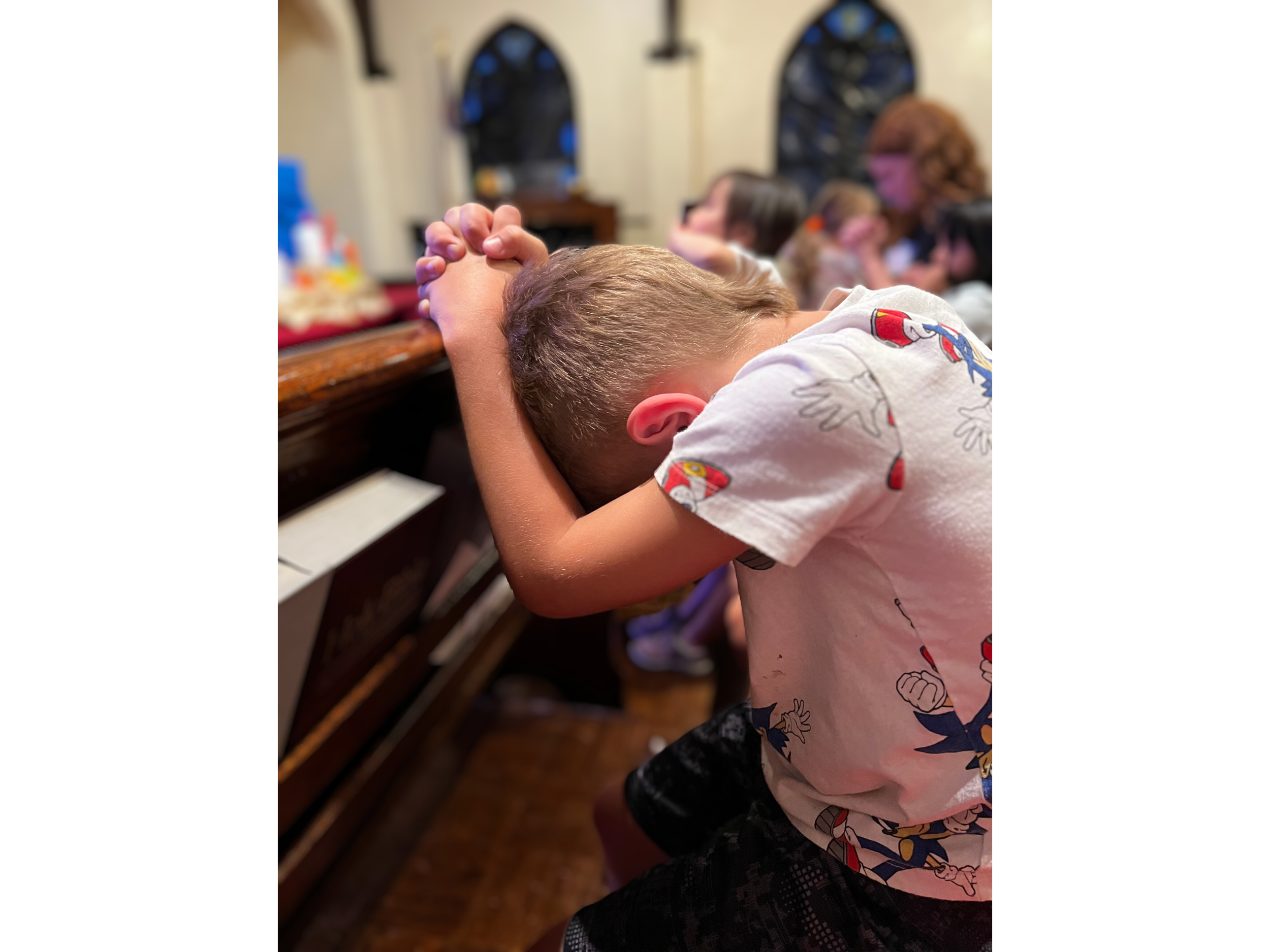 A young boy is sitting with his head bowed and hands clasped in prayer at a church or similar setting, with other children in the background also praying.