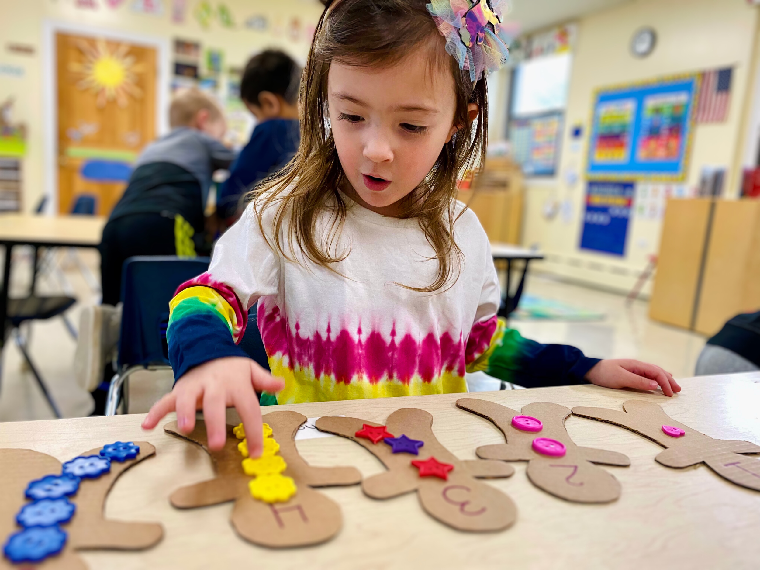 A young girl in a tie-dye shirt and a colorful bow in her hair is playing in a classroom. She is attaching colorful buttons and foam shapes onto paper reindeer cutouts on a table, which are numbered and decorated with various craft supplies.