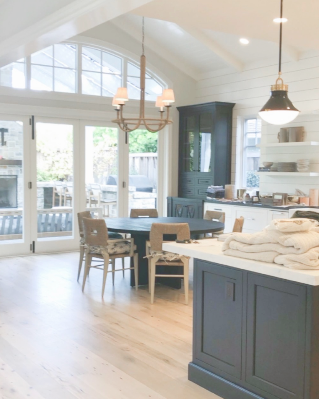 Bright open kitchen with sliding glass doors leading to outdoor patio, dark dining table with chairs, white cabinets, dark china cabinet, and pendant lighting.