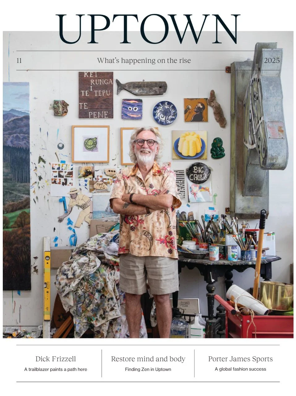A smiling man with glasses, long gray hair, and a beard, standing with his arms crossed in an artist's studio filled with colorful artwork, paintings, and art supplies on shelves and tables.