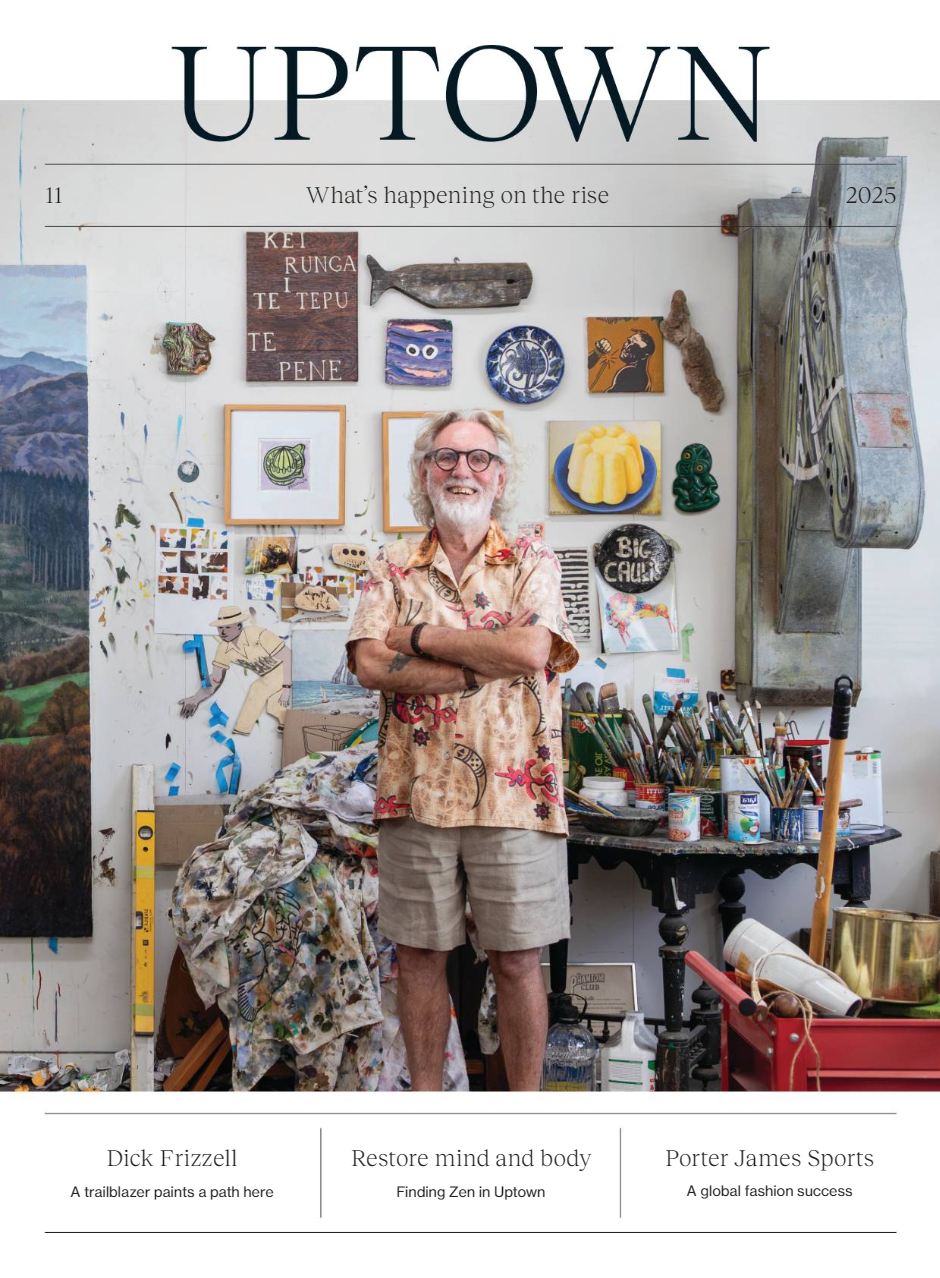 A smiling man with glasses, long gray hair, and a beard, standing with his arms crossed in an artist's studio filled with colorful artwork, paintings, and art supplies on shelves and tables.