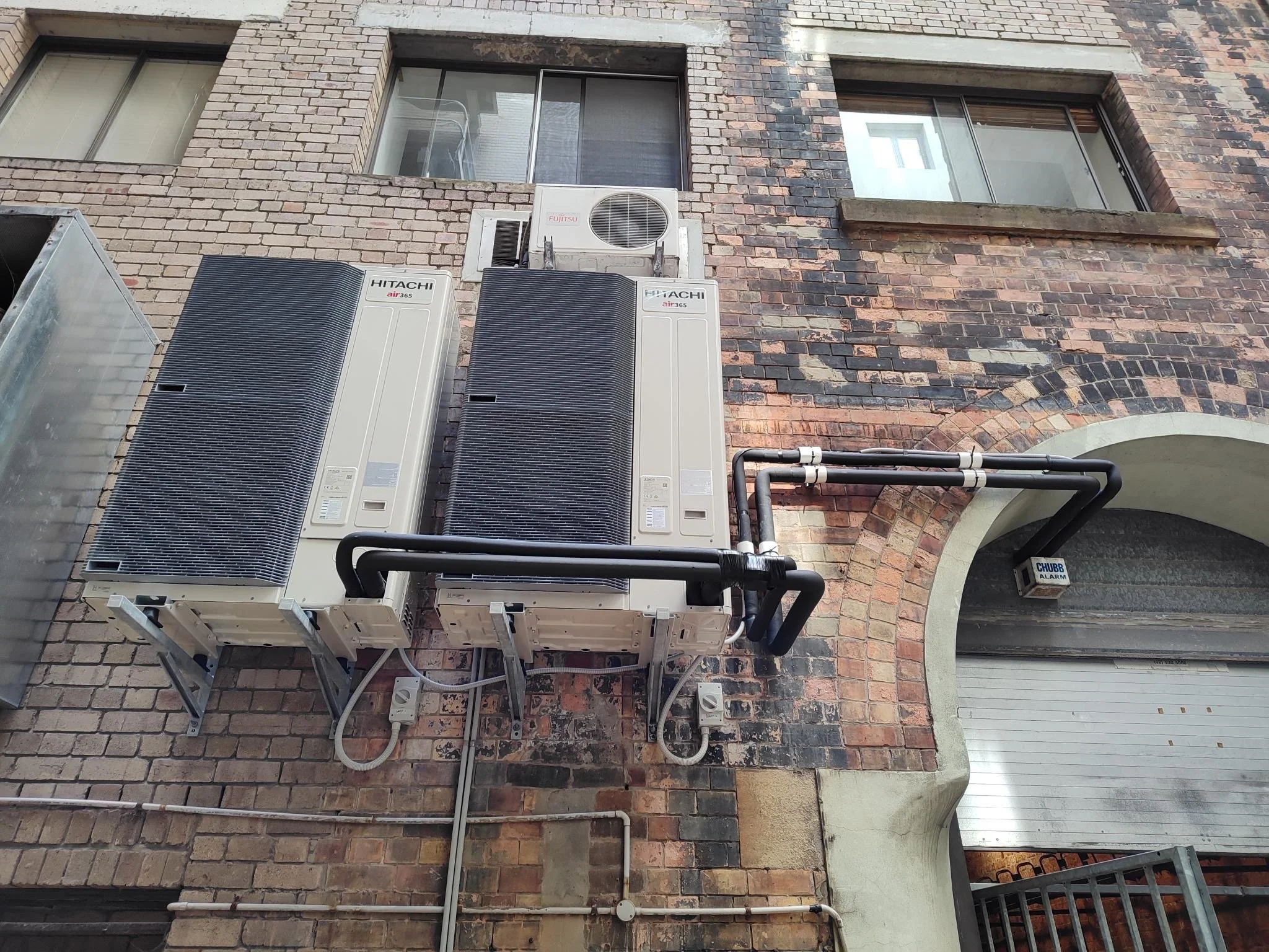 The exterior brick wall of a building with two air conditioning units and pipes attached, and a window with a blind.