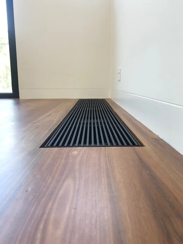 Close-up of a black floor vent on a wooden floor next to a tiled wall.