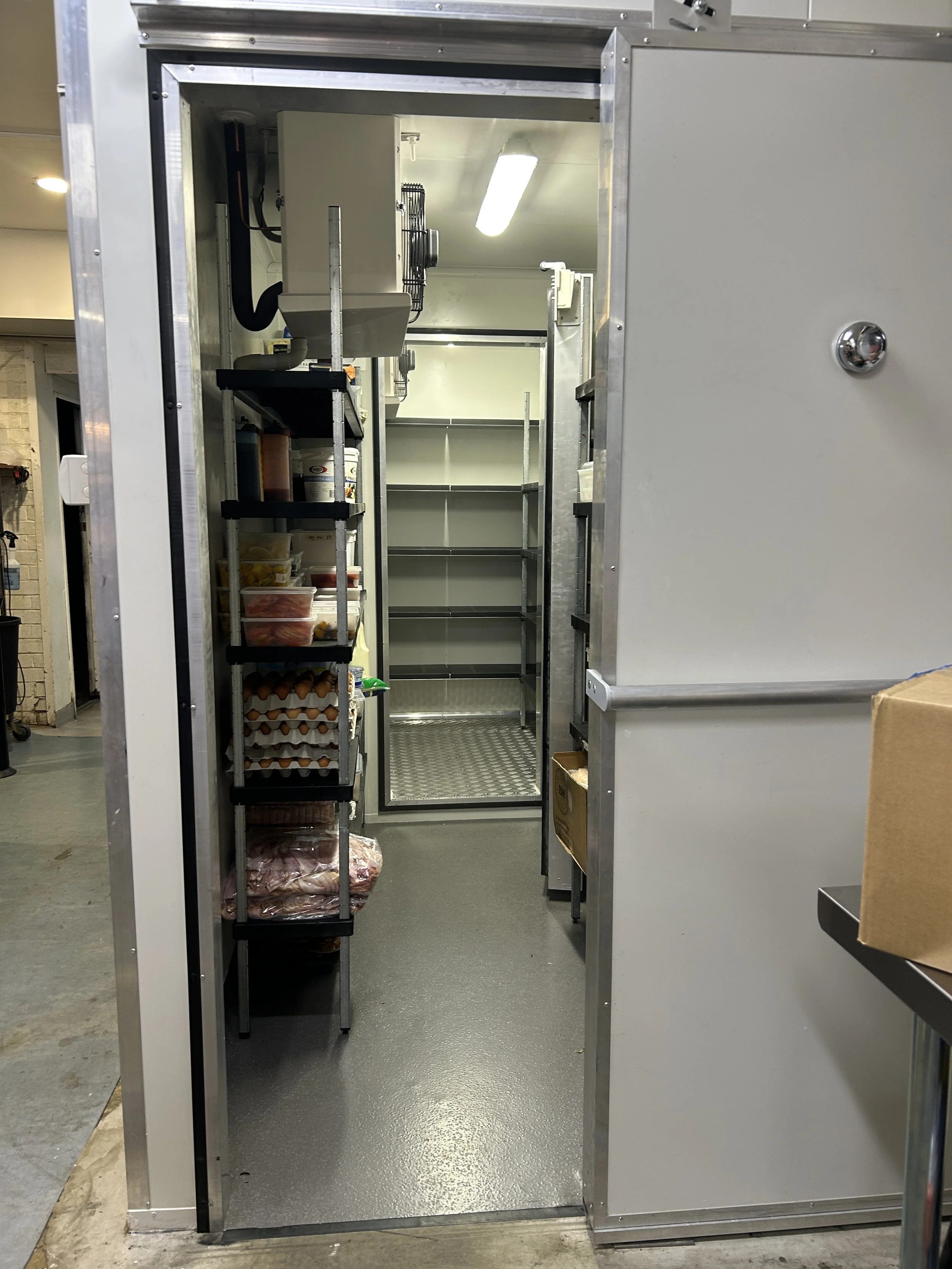 Empty walk-in refrigerator with shelves and food storage containers inside.