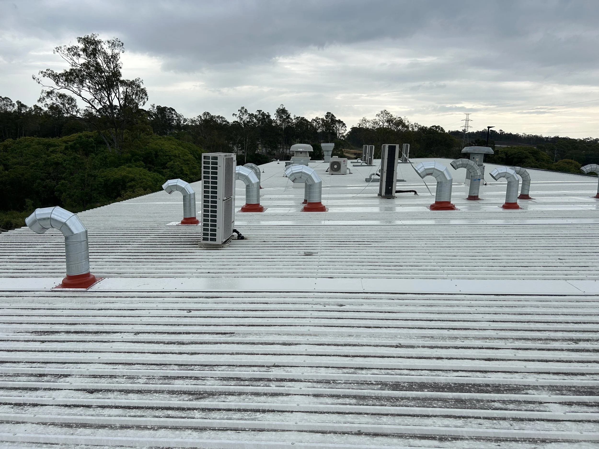 Metal roof with multiple ventilation pipes and air conditioning units, with trees and cloudy sky in the background.
