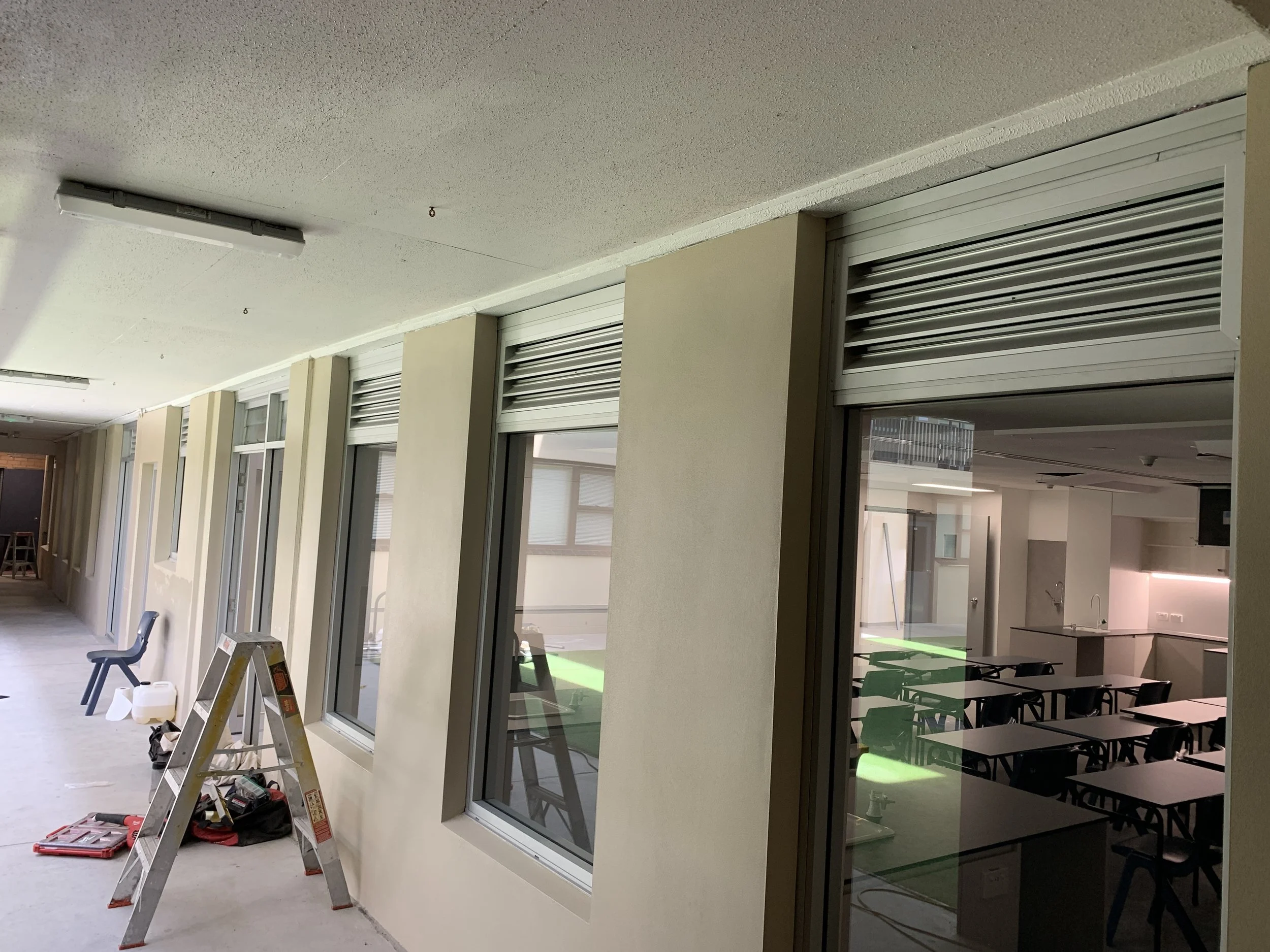 A corridor with large windows on the left side and a classroom or office space with tables and chairs visible through a partially open rolling shutter on the right. Construction tools and a ladder are on the floor.