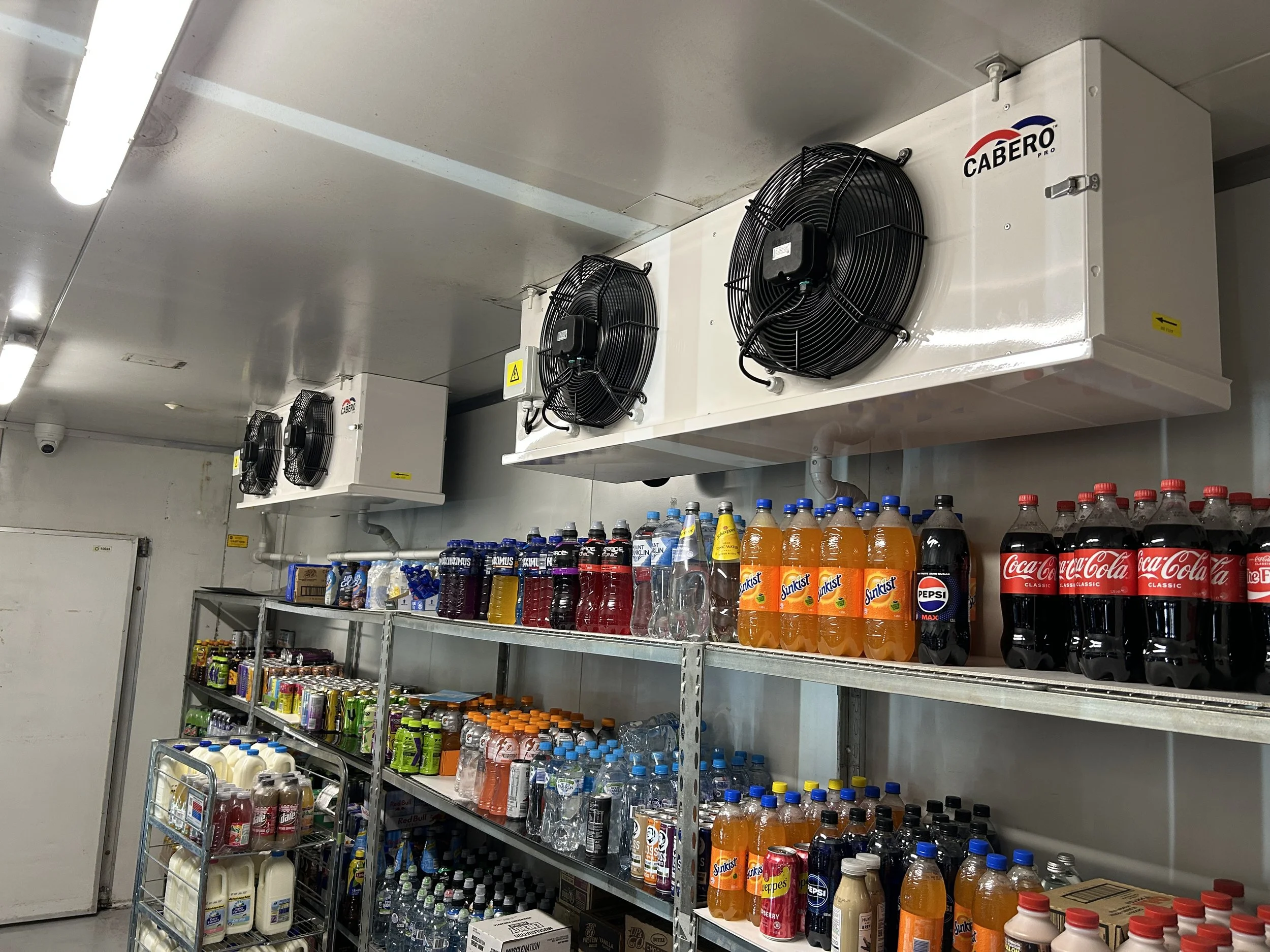 Shelves stocked with various bottled drinks including sodas, juices, and water bottles, under industrial cooling units with fans. Port Macquarie, New South Wales. Refrigeration Services