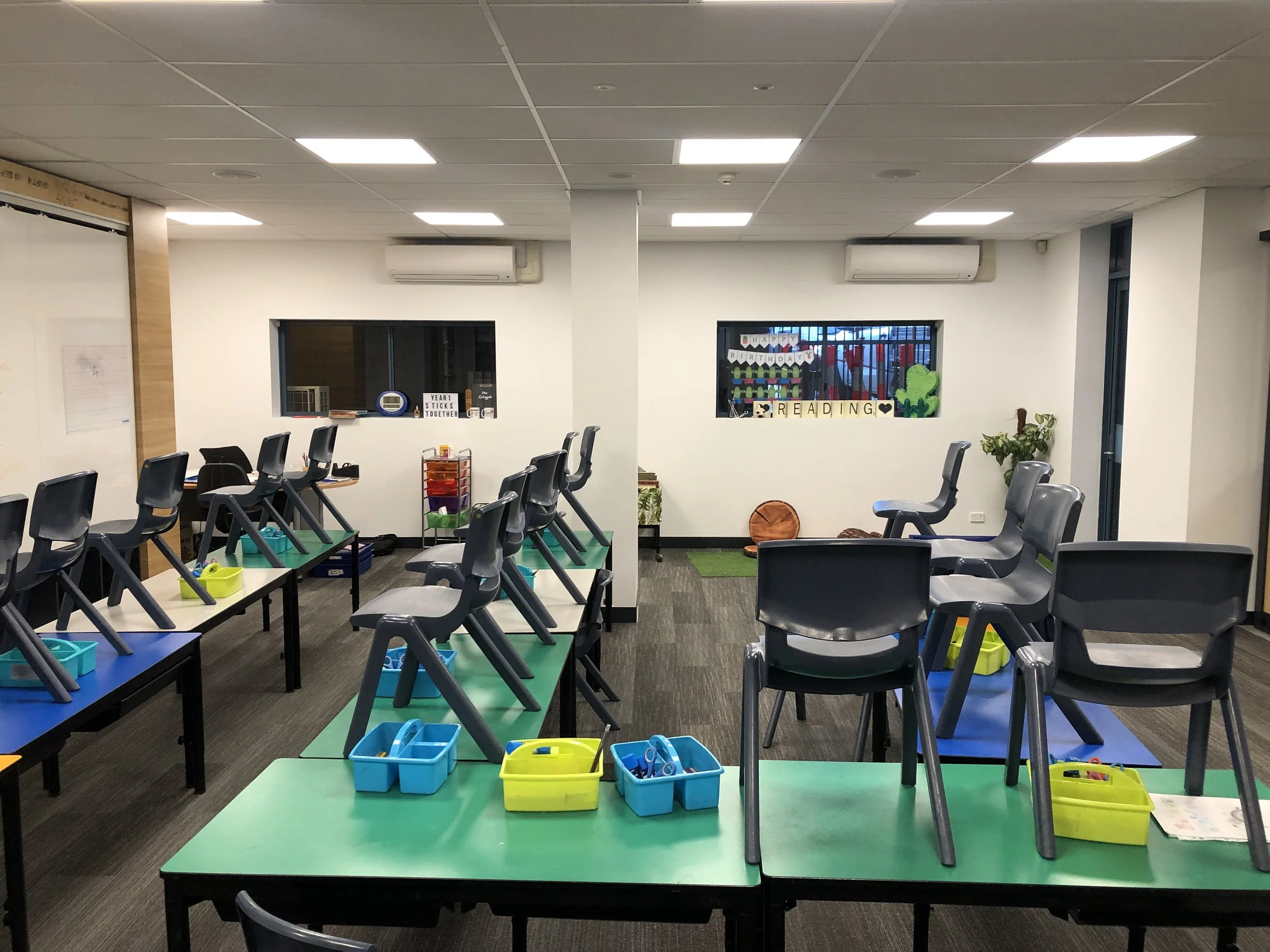 Children's classroom with chairs placed upside down on tables, colorful bins on tables, and educational decorations and books on the wall.