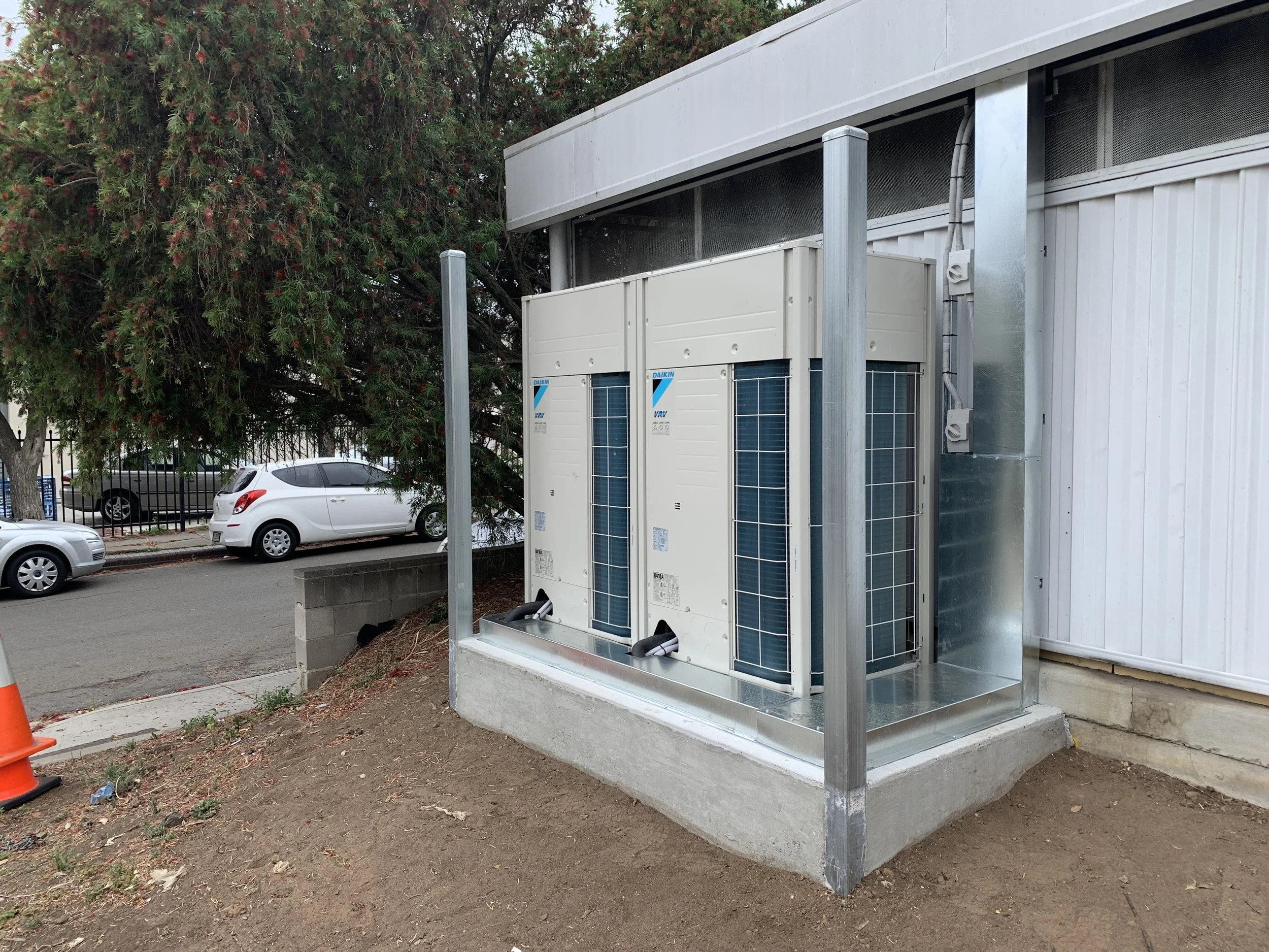 Outdoor view of HVAC units installed on a concrete pad beside a building with trees and parked cars in the background.