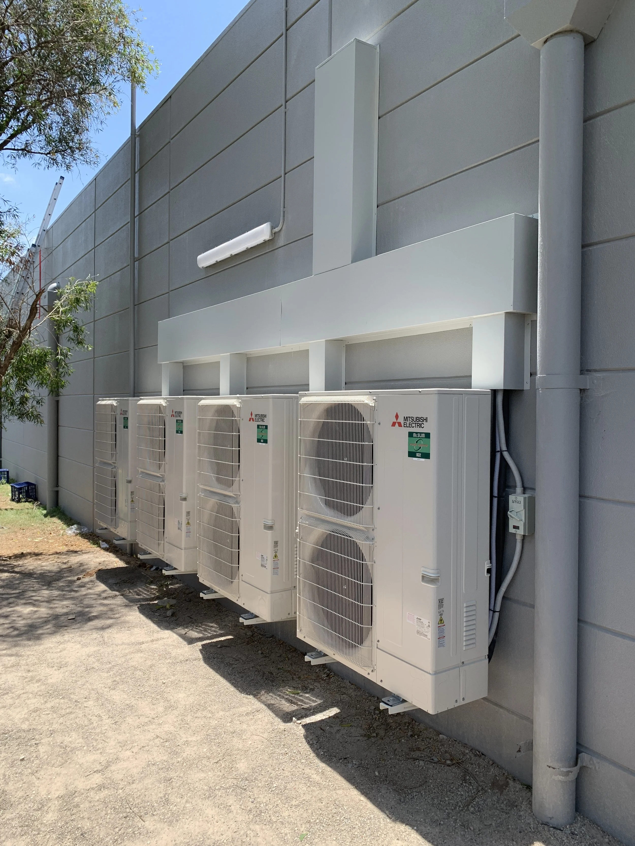 Four Mitsubishi electric air conditioning outdoor units installed on a concrete wall outside a building, with electrical wiring and a conduit, under a clear blue sky.