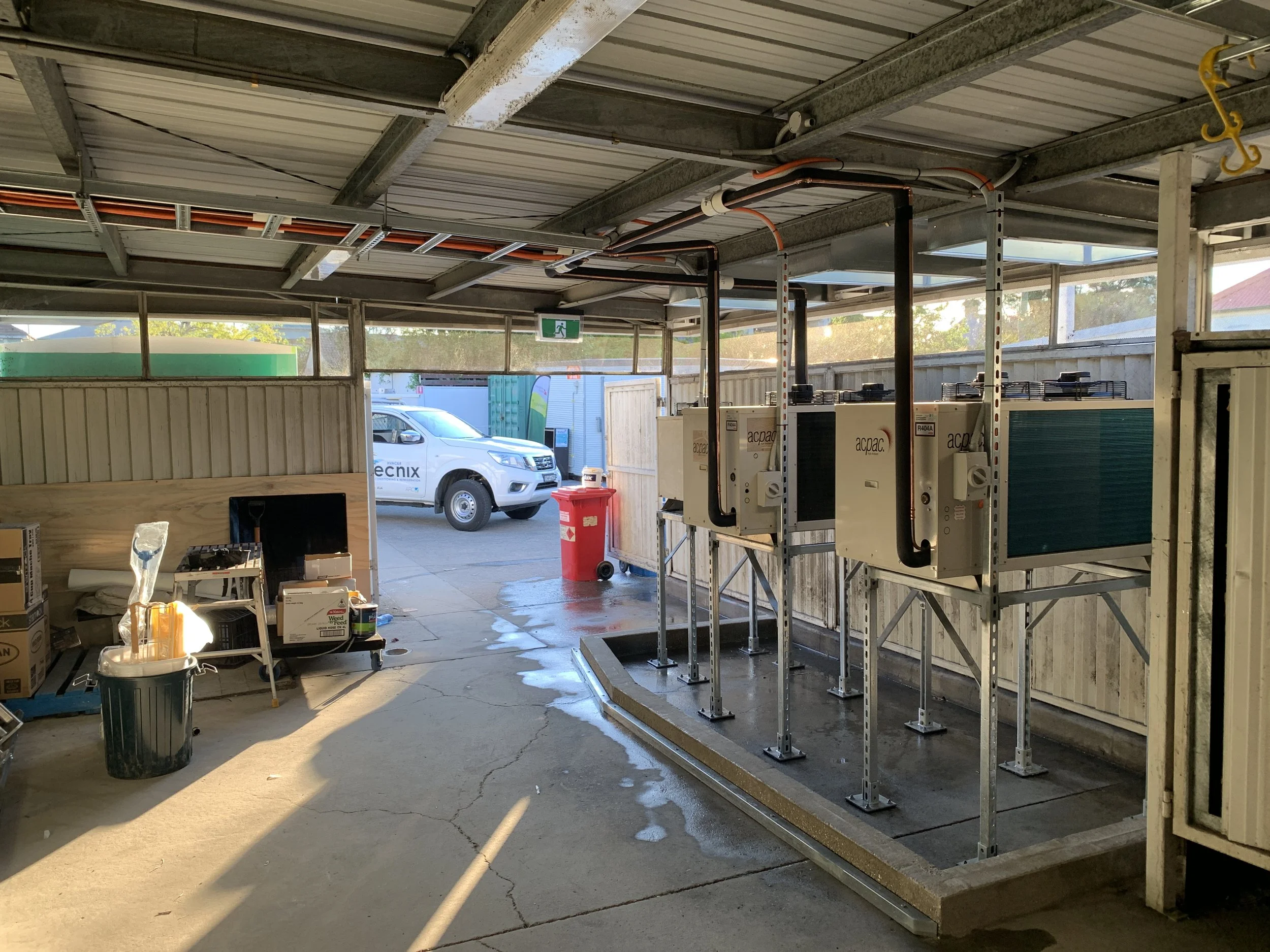 View of an industrial or commercial garage with electrical or mechanical equipment, a parked white vehicle outside, some boxes, and a focus on plumbing or HVAC units.