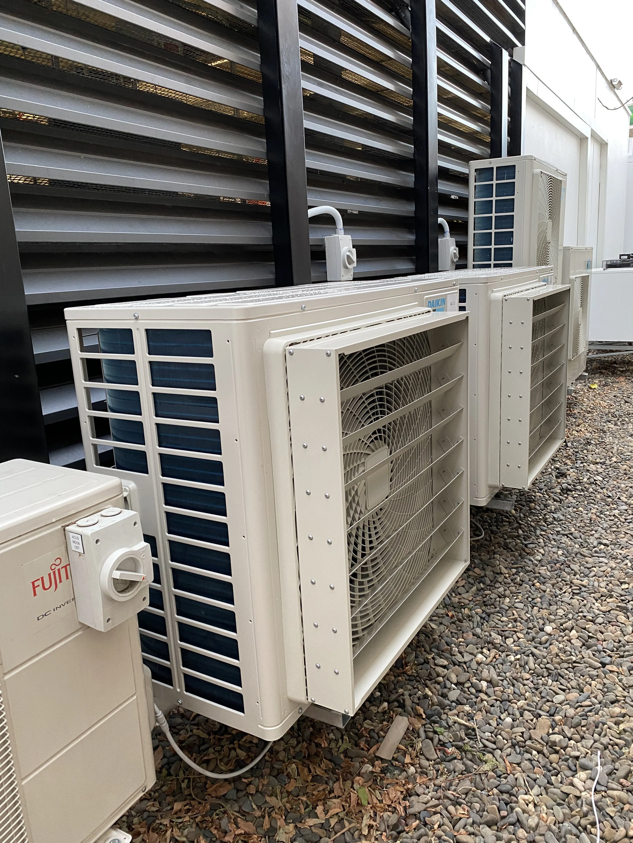 Several air conditioning units installed outdoors next to a building, with a gravel ground and a black metal wall in the background.