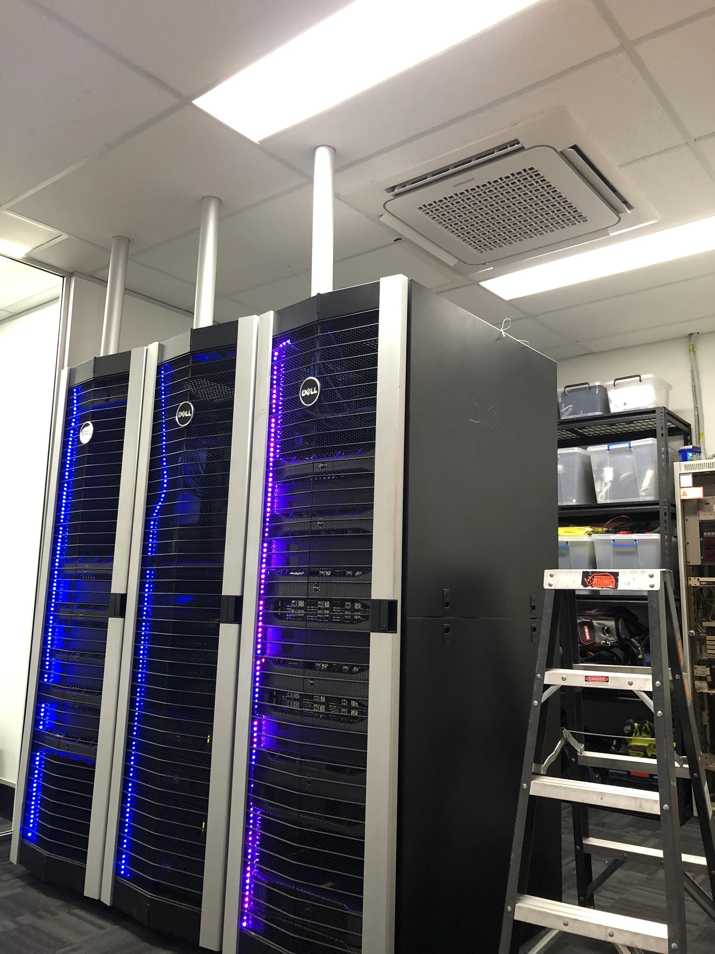 Server room with three Dell server racks, black and gray, with blue and purple LED lights, and a small ladder on the right side. In the background, shelves with storage bins and tools.