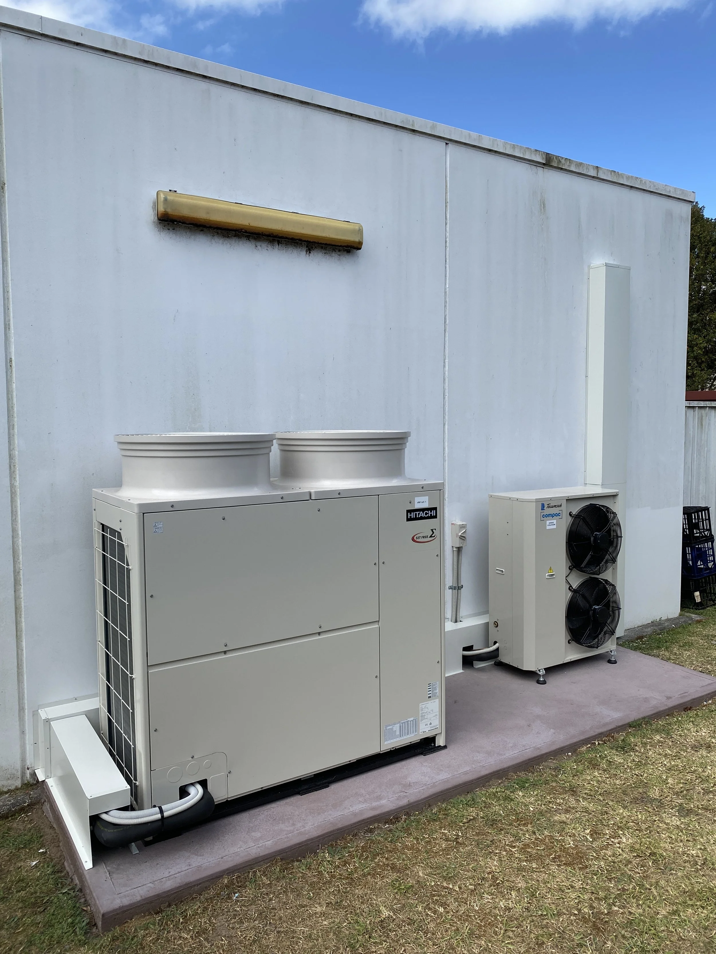 Outdoor HVAC units, including a larger heat pump and a smaller condensing unit, installed on a concrete pad against a white wall, with a blue sky above.