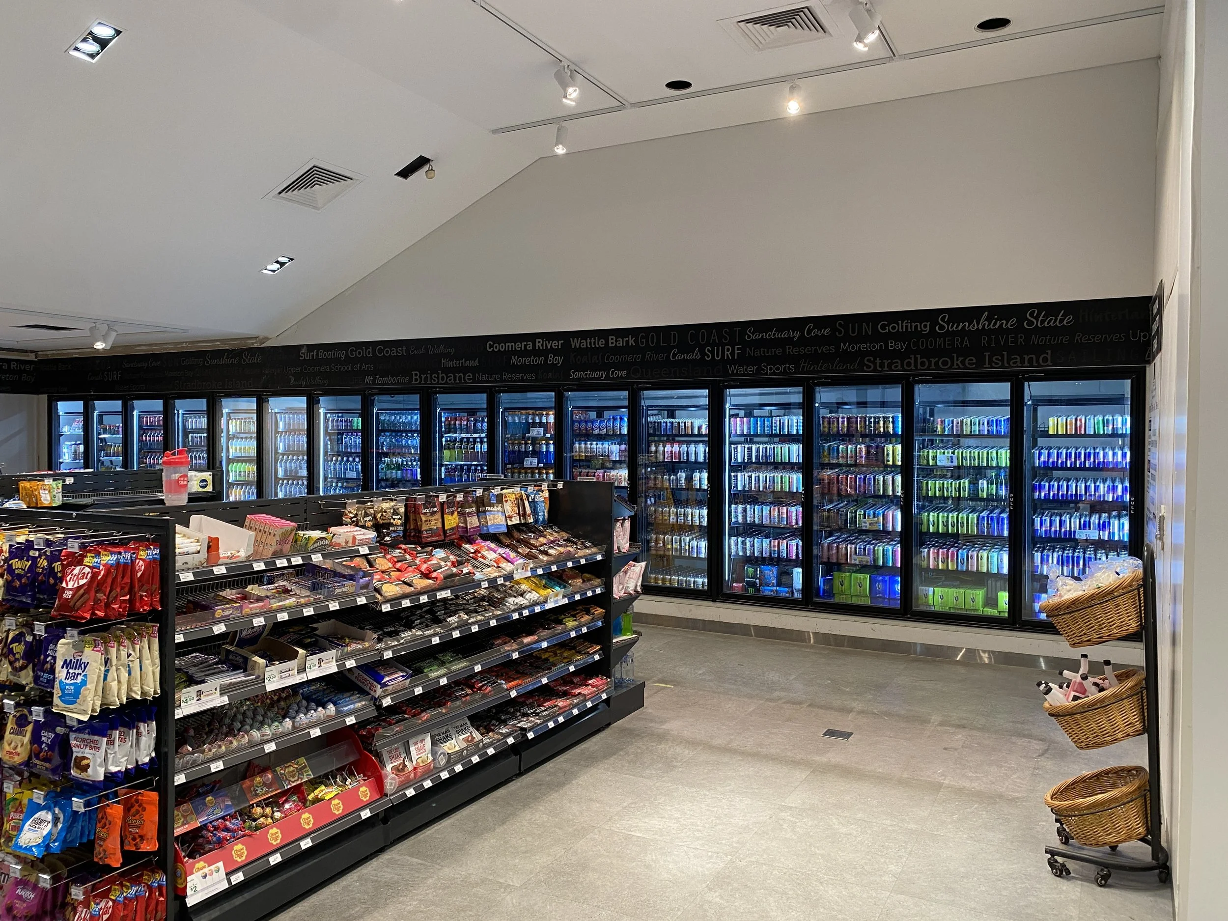 Supermarket aisle with refrigerated drinks and snack shelves, including chips, chocolates, and candies, and wicker baskets on a rolling cart on the right.
