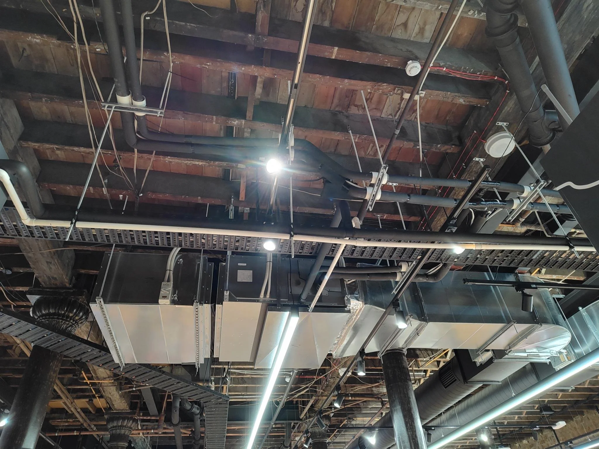 Ceiling view of industrial HVAC system with ducts, pipes, and lighting in a commercial or office building.