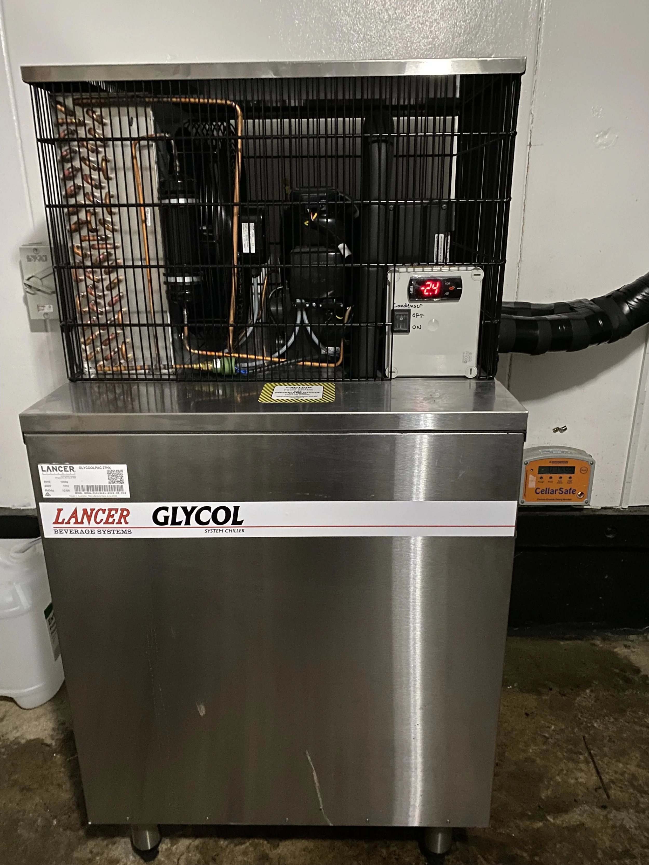 Commercial glycol chiller unit with a metal exterior, black wire cage on top, digital temperature display reading -24°C, and an orange safety device on wall nearby.
