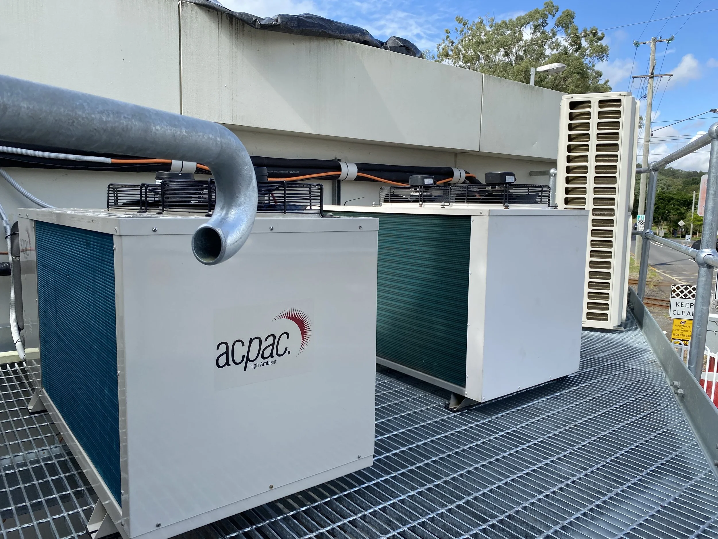 Three large air conditioning units on a metal platform outside a building, with pipes and electrical wiring connecting them. Sunshine Coast, Queensland