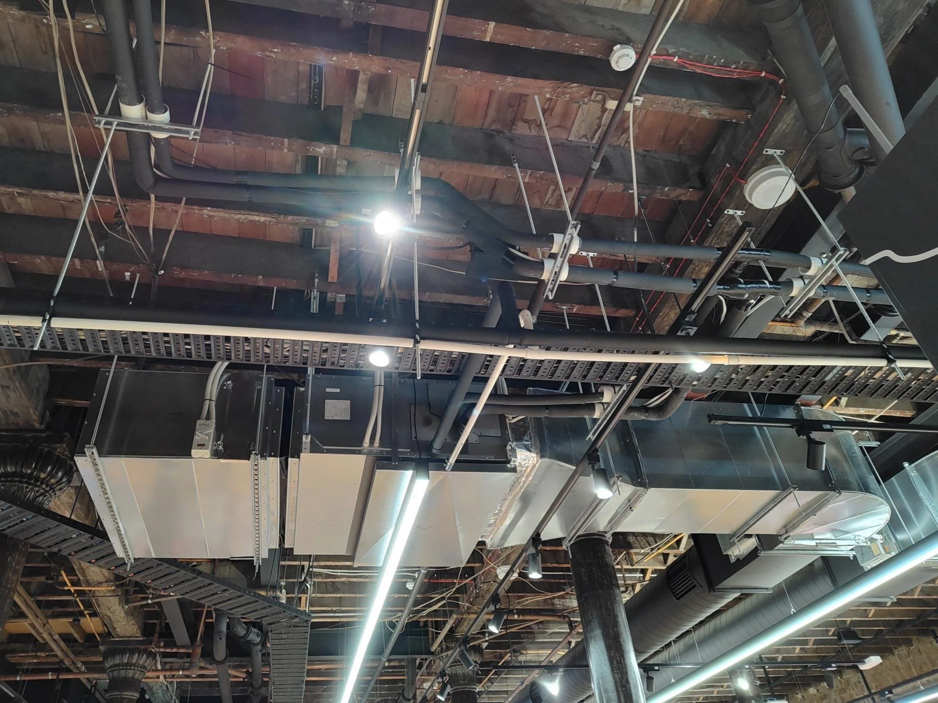 Ceiling of an industrial building with exposed pipes, ventilation ducts, electrical wiring, and lighting fixtures.
