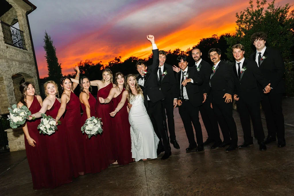 Group of wedding party members, including bridesmaids in red dresses, a bride in a white gown, and groomsmen in black suits, posing outdoors at sunset with colorful sky in background.