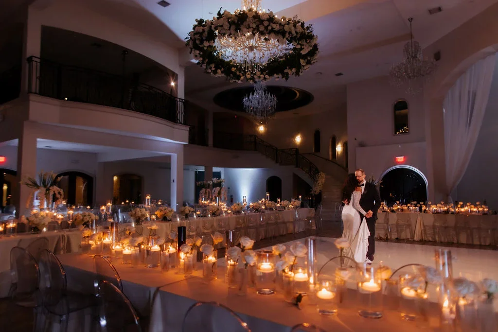 A couple is dancing on a decorated wedding reception dance floor with candles, flowers, and elegant lighting in a grand hall.