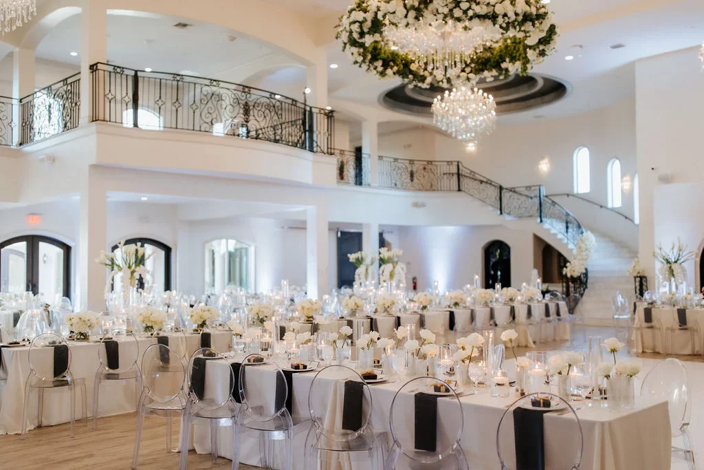 Elegant event hall decorated for a wedding reception with long tables covered in white tablecloths, floral centerpieces, candles, and clear ghost chairs, featuring a high ceiling adorned with crystal chandeliers and a curved staircase.