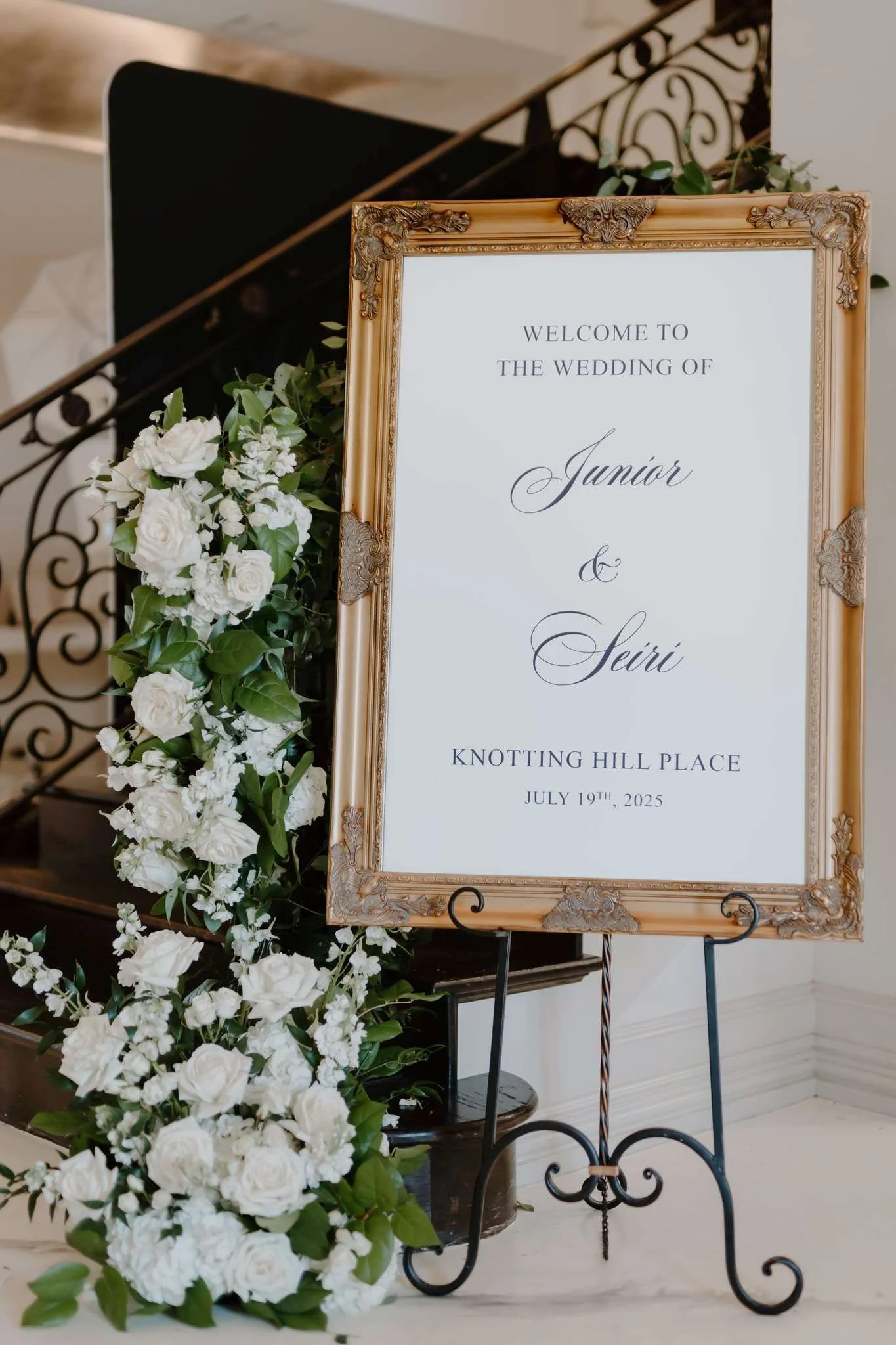 Elegant wedding welcome sign framed in gold with white roses and green foliage decoration, indicating the wedding of Junior and Seiri on July 19th, 2025, at Knotting Hill Place.