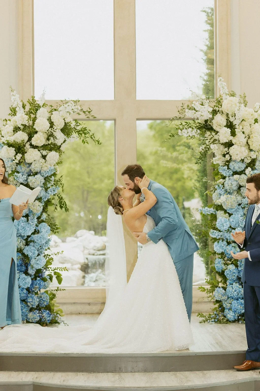 A bride and groom standing on a stage, kissing during their wedding ceremony, with an officiant and two groomsmen present, floral arrangements in the background.