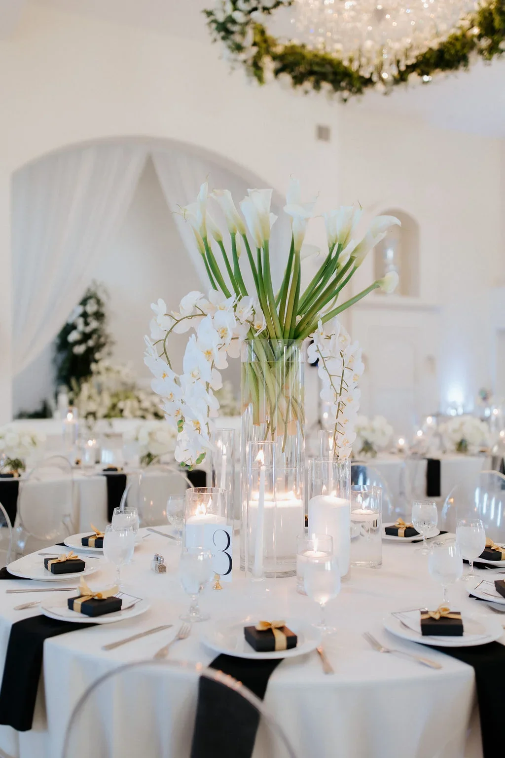 Elegant wedding reception table with a tall glass vase filled with white calla lilies and orchids, surrounded by lit candles, white and black table settings, and decorative black napkins with gold ribbons.