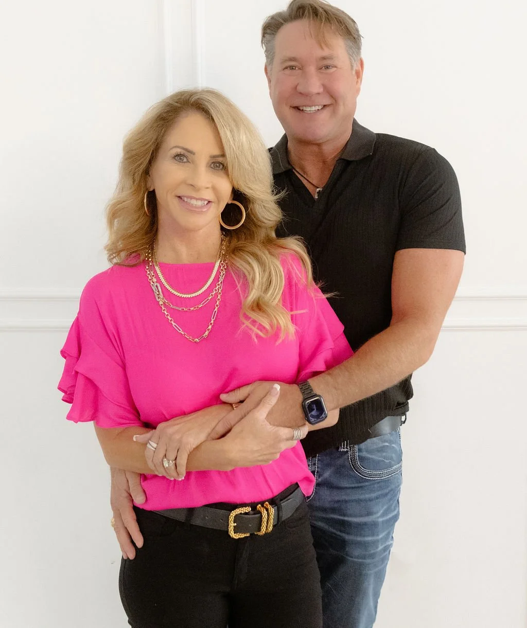 A smiling woman with long blonde hair, wearing a bright pink blouse with ruffled sleeves, gold jewelry, and a black belt, standing in front of a man with gray hair who is wearing a black polo shirt and jeans, with both posing against a plain white wall.