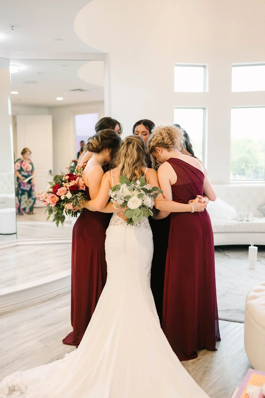 A bride and her bridesmaids share a hug at a wedding, holding bouquets of flowers, inside a bright room with large windows.