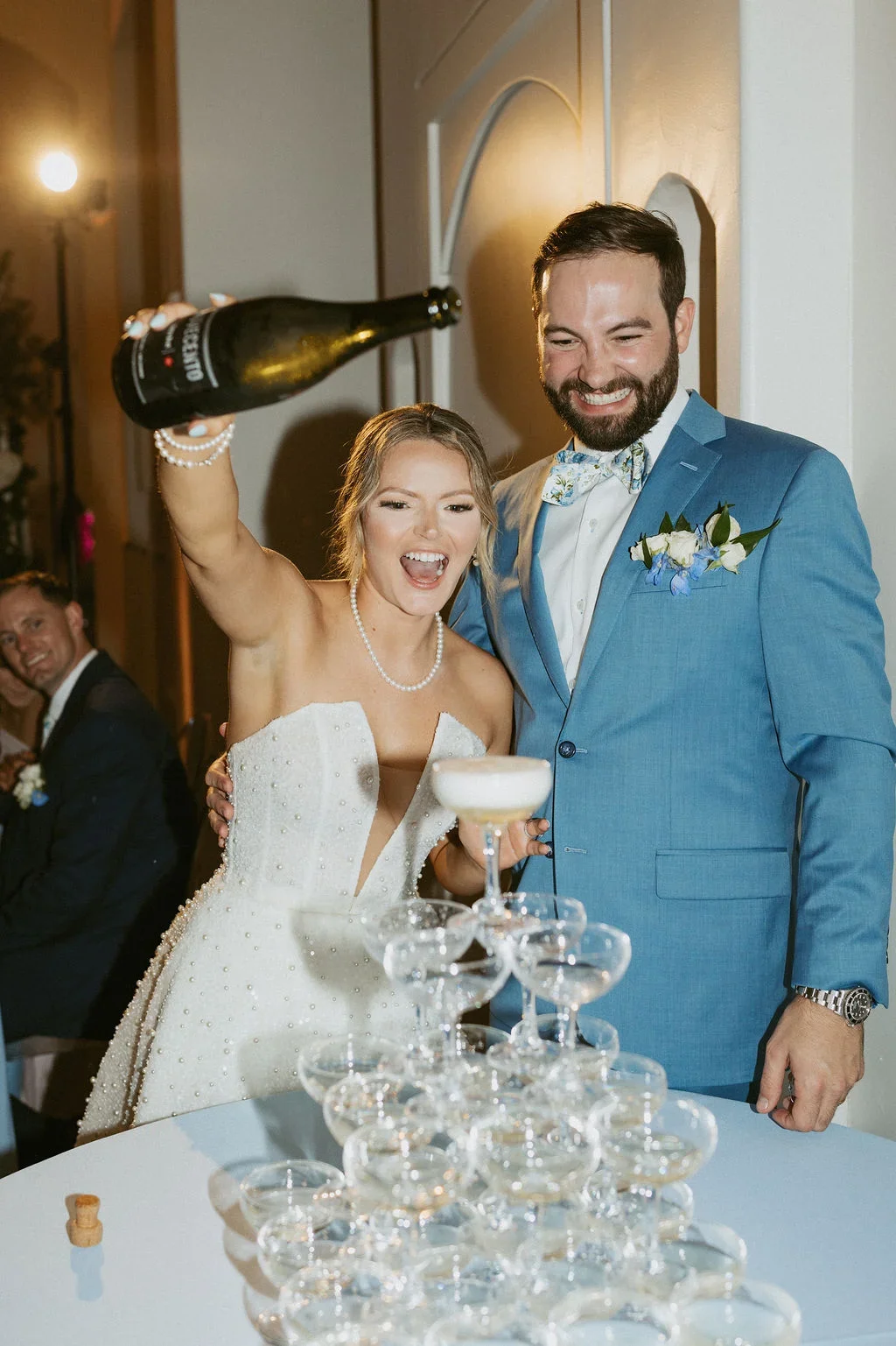 A bride and groom celebrating at a wedding reception, with the bride pouring champagne into a tower of glasses, both smiling joyfully.