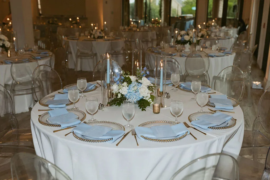 A round banquet table set for a formal event with a white tablecloth, light blue napkins, clear glass plates with pearl-like rims, water glasses, and gold utensils. The centerpiece features tall glass candle holders with white candles, blue and white
